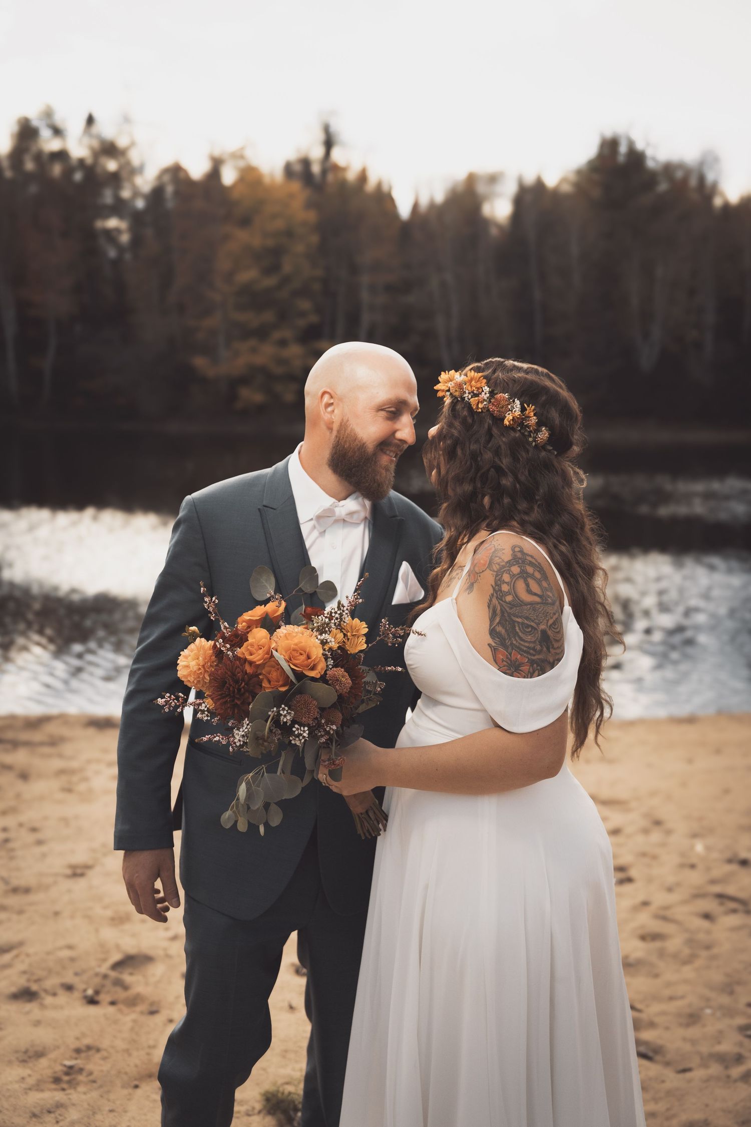 A romantic autumn wedding couple stands on a beach by a lake holding a bouquet of orange flowers at sunset.