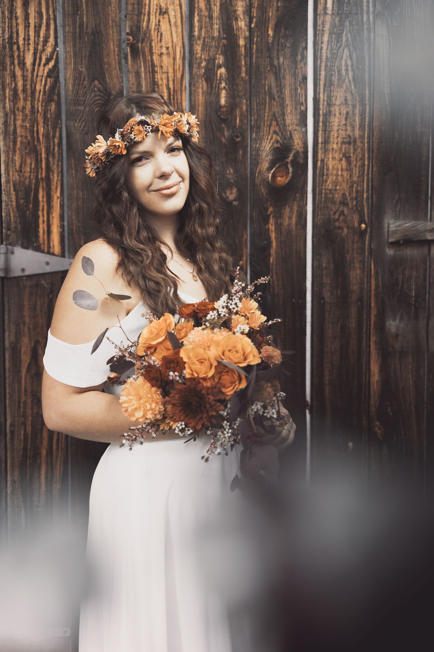 Bride with floral crown and bouquet poses against rustic wooden wall.