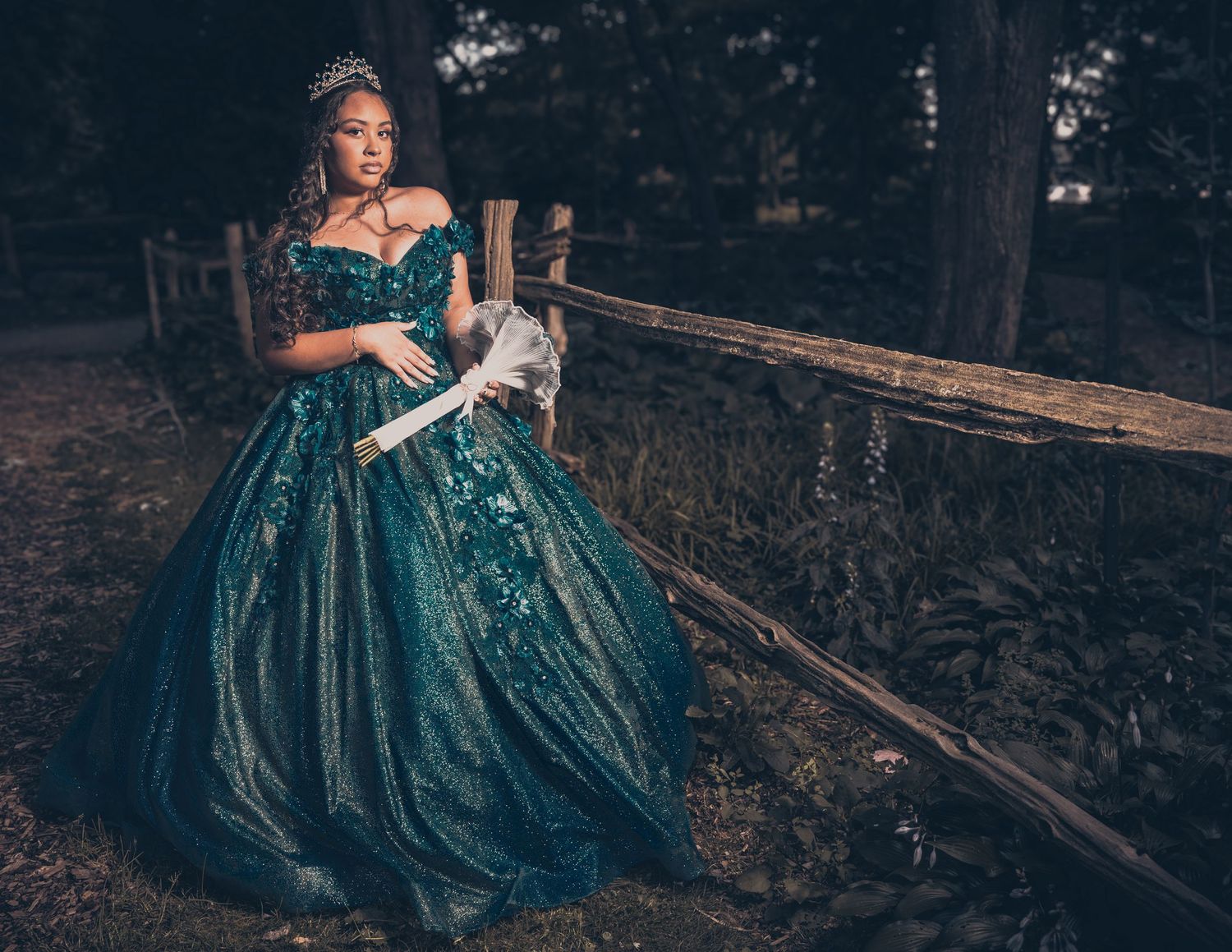 Person in teal ball gown poses near rustic wooden fence at dusk.