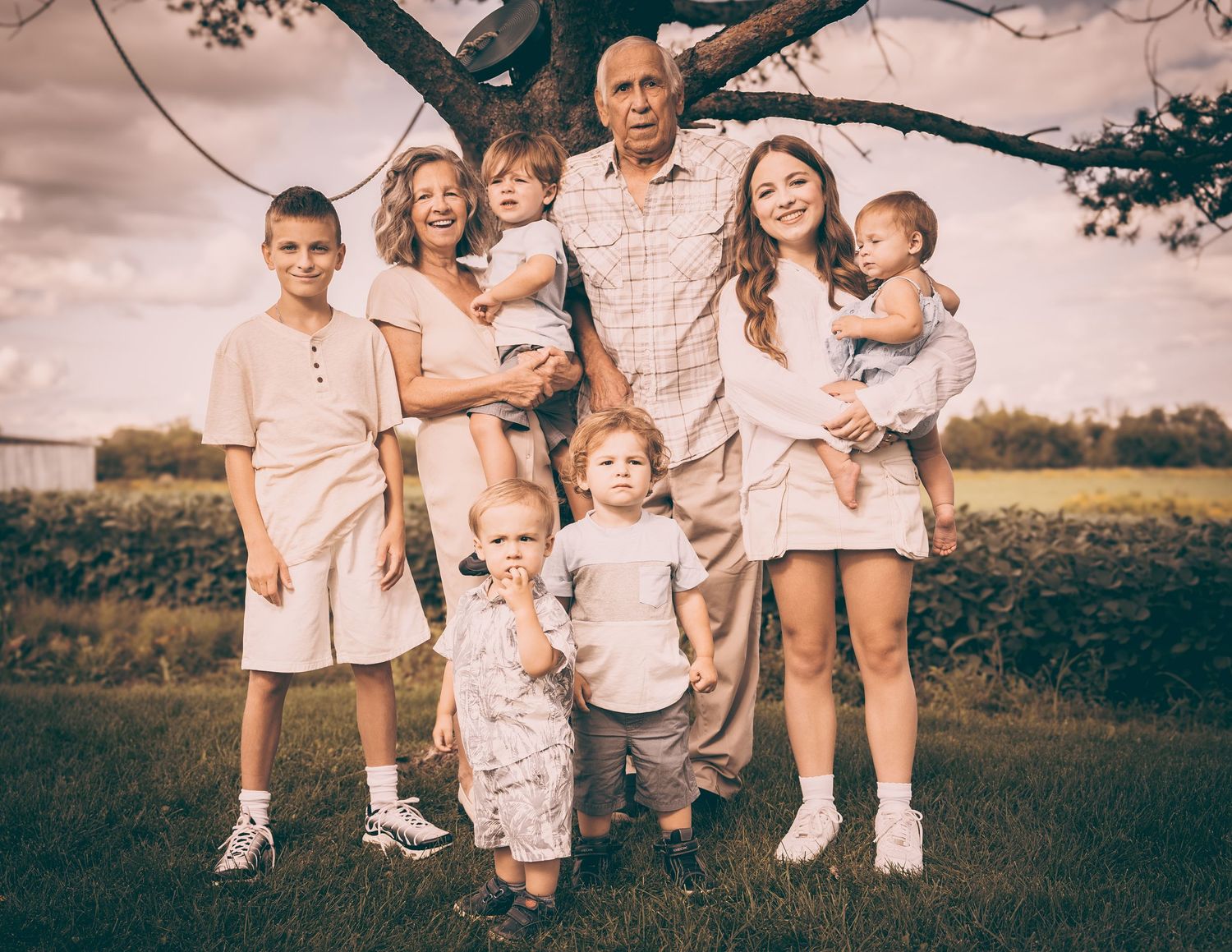 Multi-generational family portrait outdoors under a tree in soft vintage lighting.