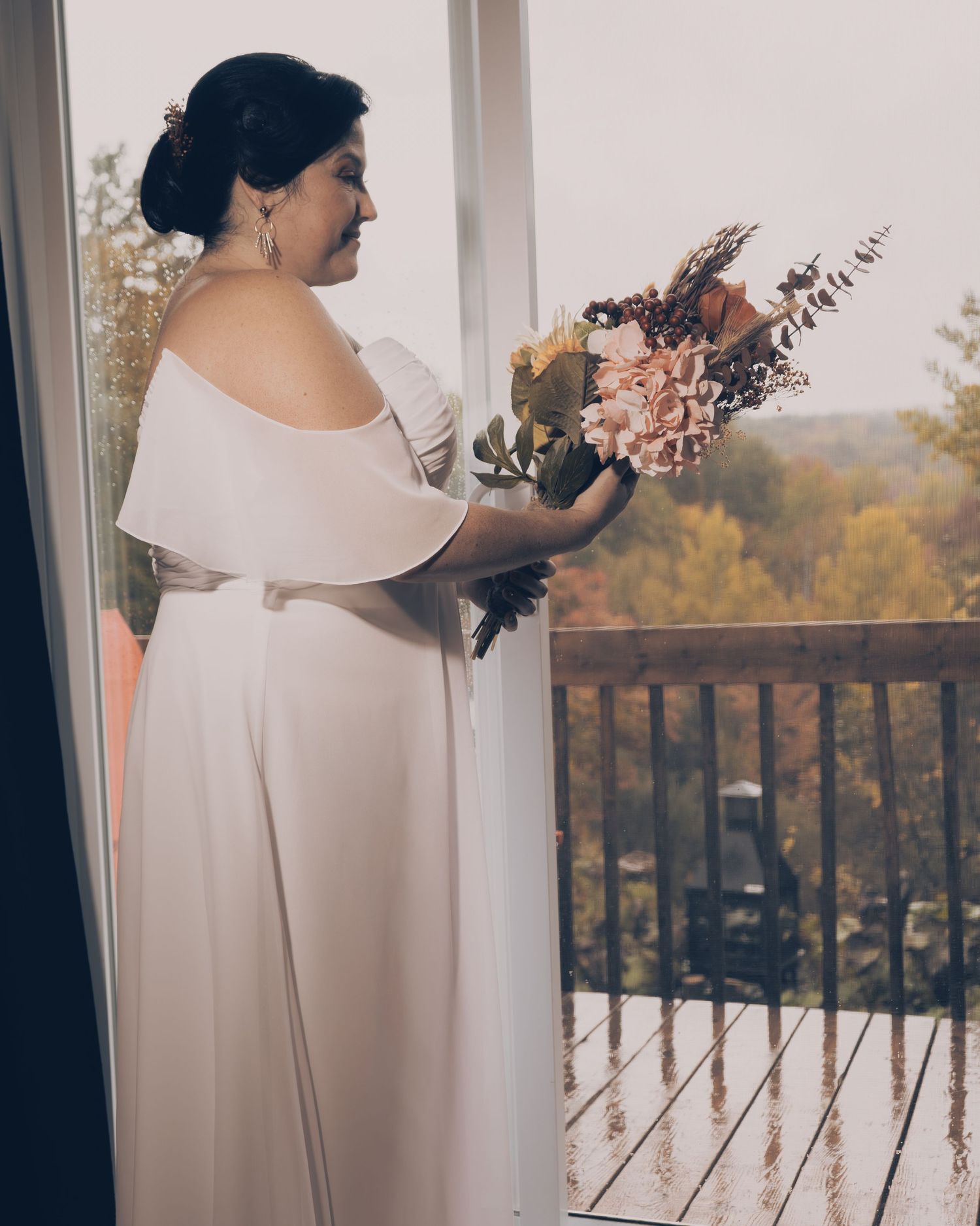 Figure in an elegant off-shoulder white gown holds a muted-toned dried floral bouquet while standing on a balcony at sunset.