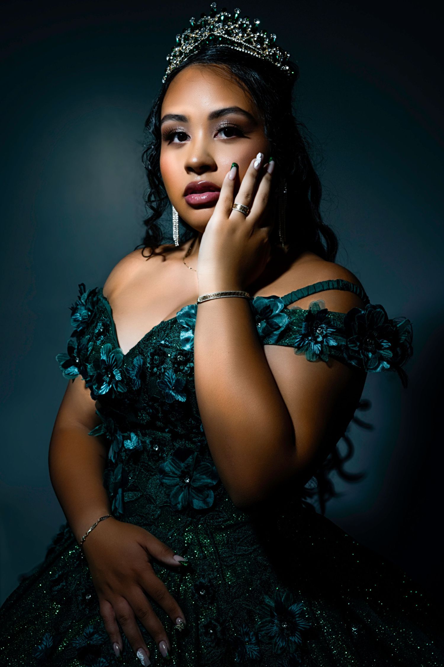 Dramatic portrait shot of a quinceañera in a sparkly teal ball gown and tiara against a dark background.