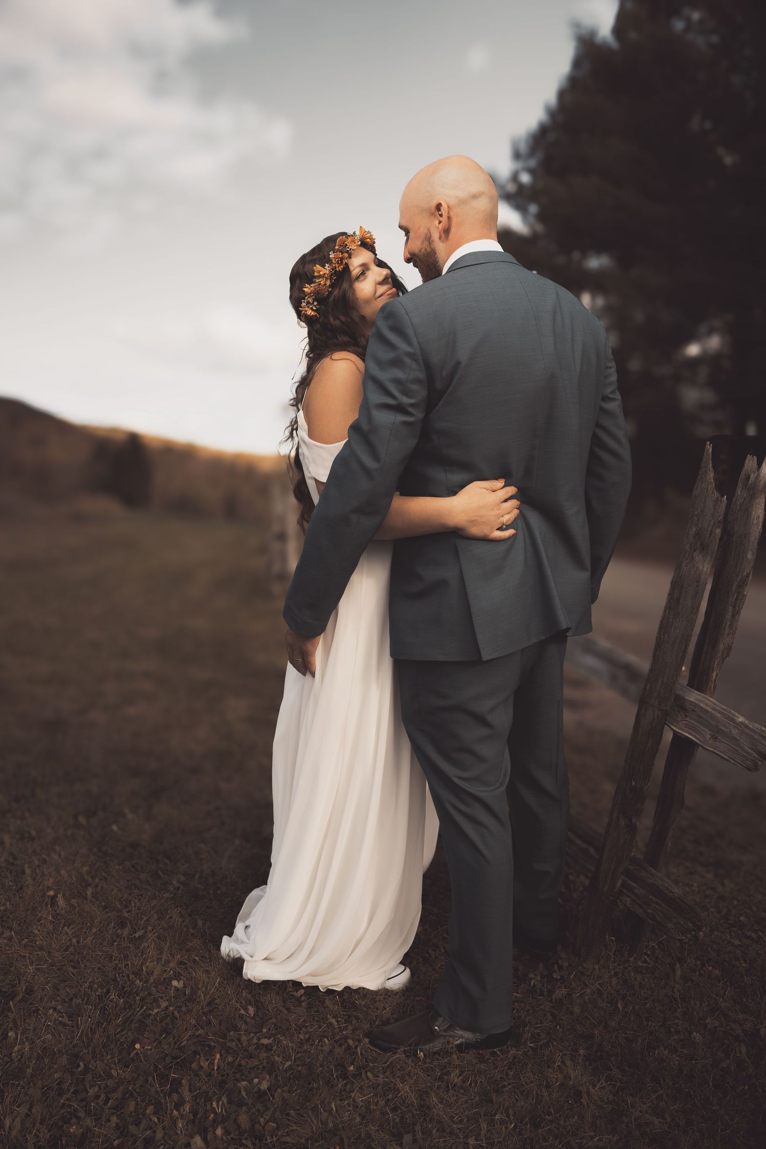 A romantic wedding couple embraces outdoors against a moody sky, with the bride in white and groom in gray suit.