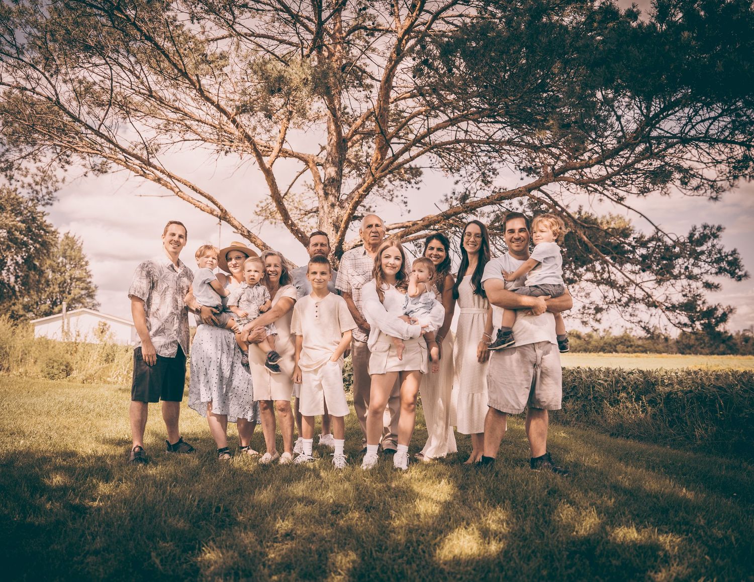 A casual family group photo outdoors wearing coordinated white and beige outfits under a large tree.
