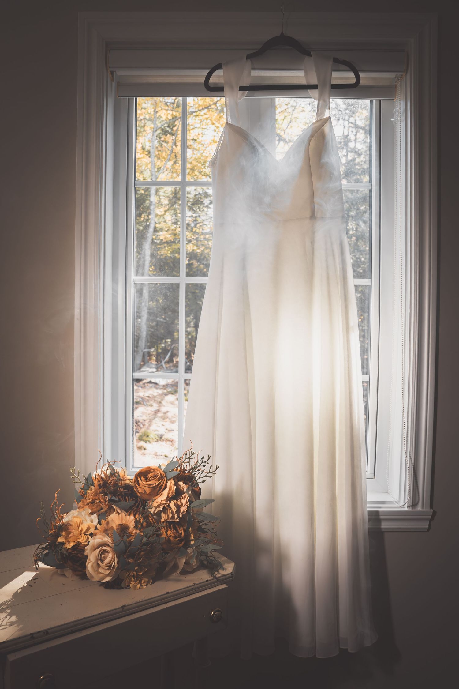 White wedding dress hanging in window with dried orange floral bouquet.