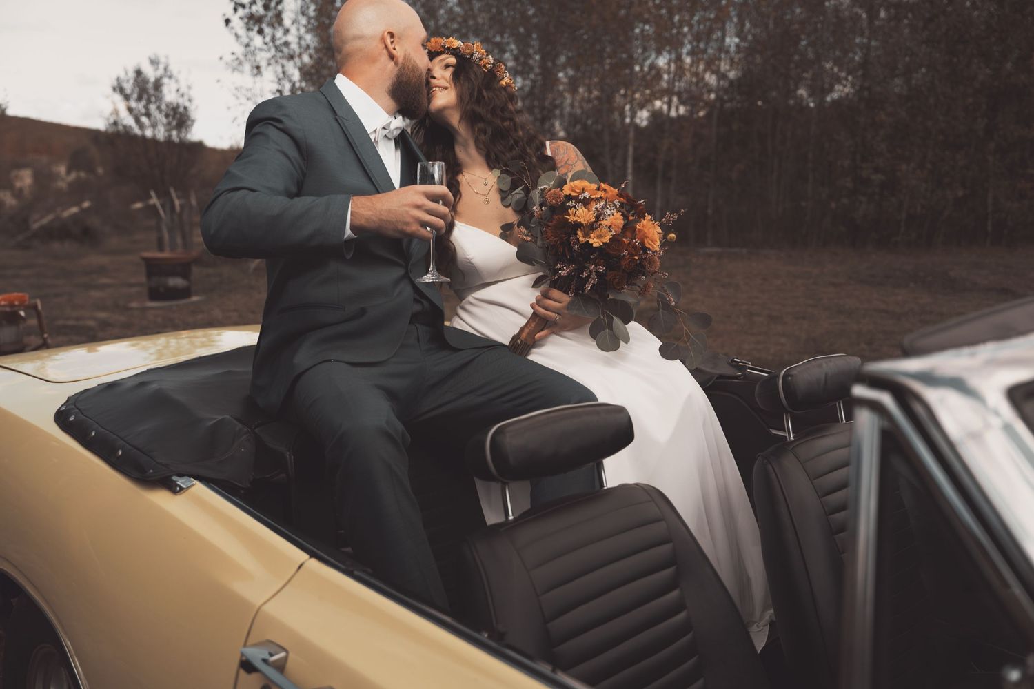 A couple in wedding attire sits together on a vintage tan-colored Vespa scooter holding a fall-themed bouquet.