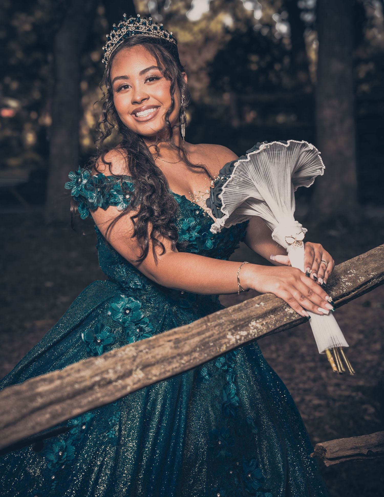 Person in teal ball gown holds white flowers while leaning on wooden railing outdoors.