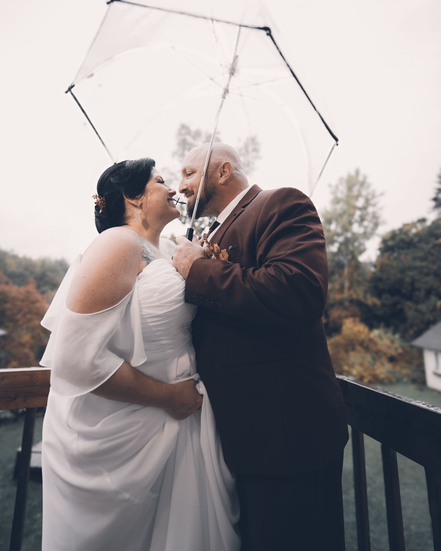 A romantic moment shared under a white umbrella on a deck surrounded by autumn foliage during a wedding celebration.