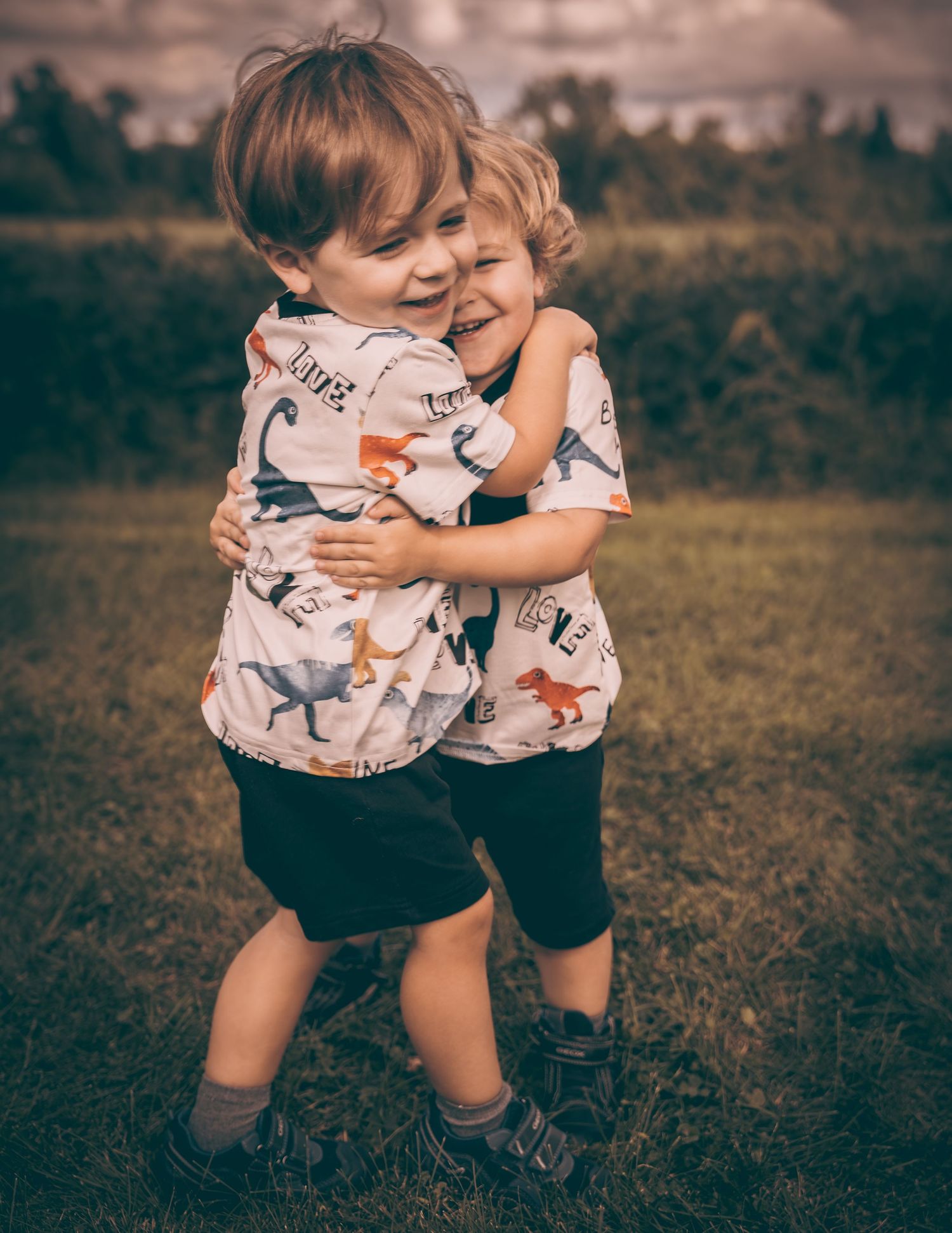 Two young children in matching dinosaur shirts share a sweet hug outdoors.