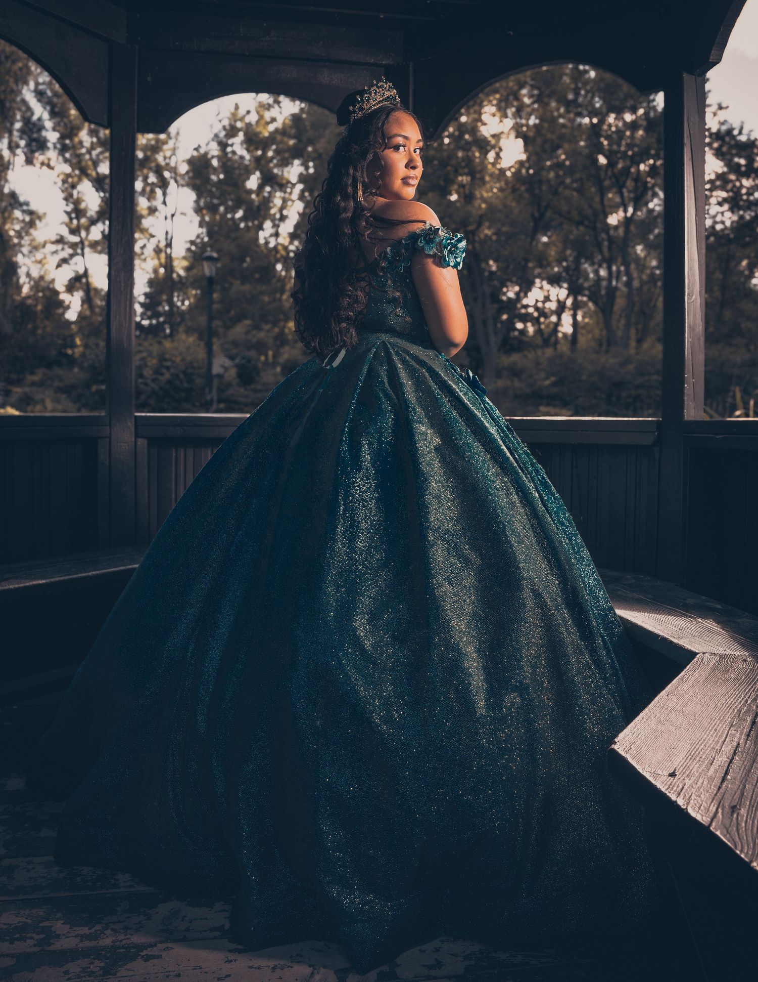 Silhouetted figure in teal ball gown poses against sunset on covered porch.