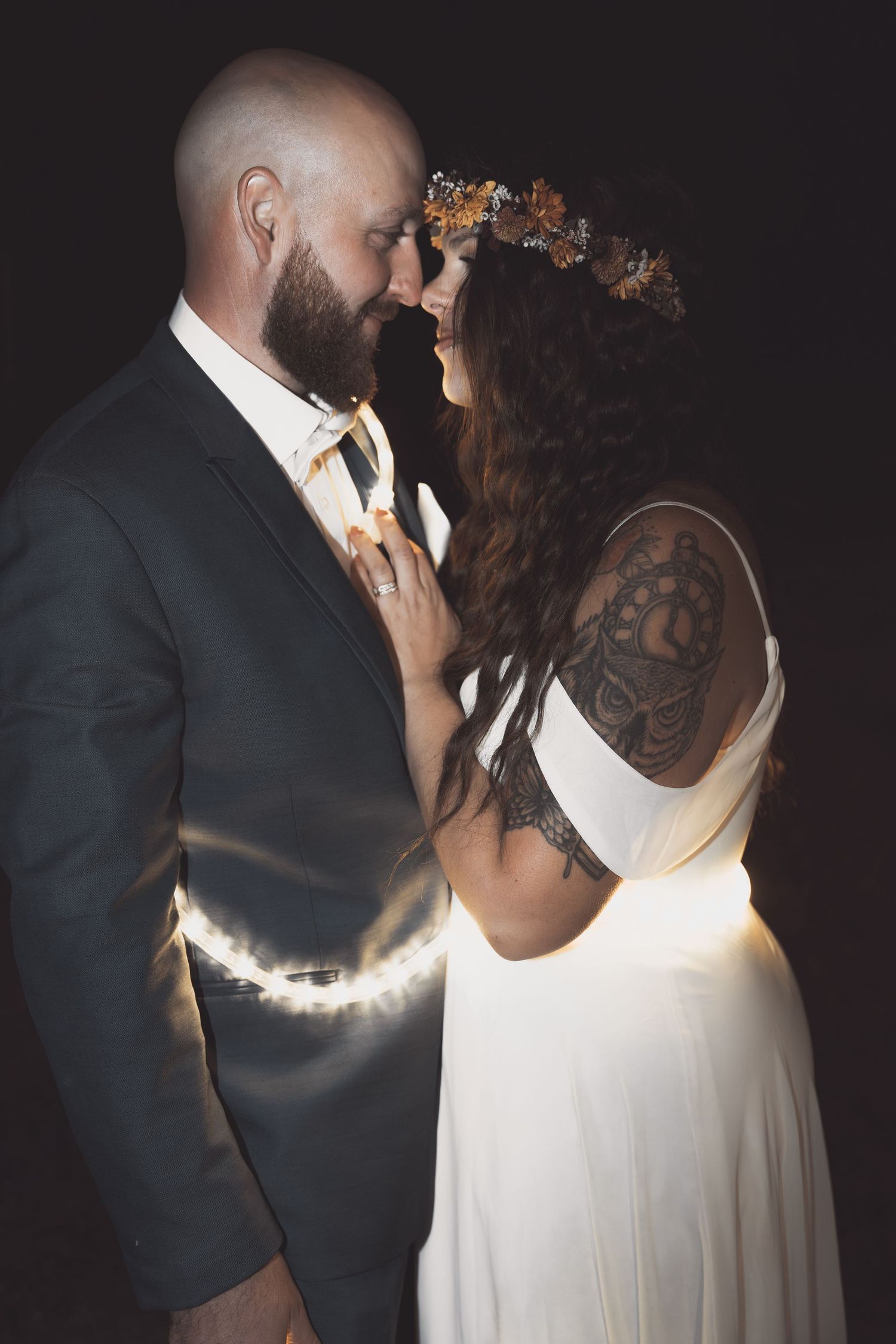 A couple in wedding attire share an intimate moment with dramatic backlit effect against a dark background.