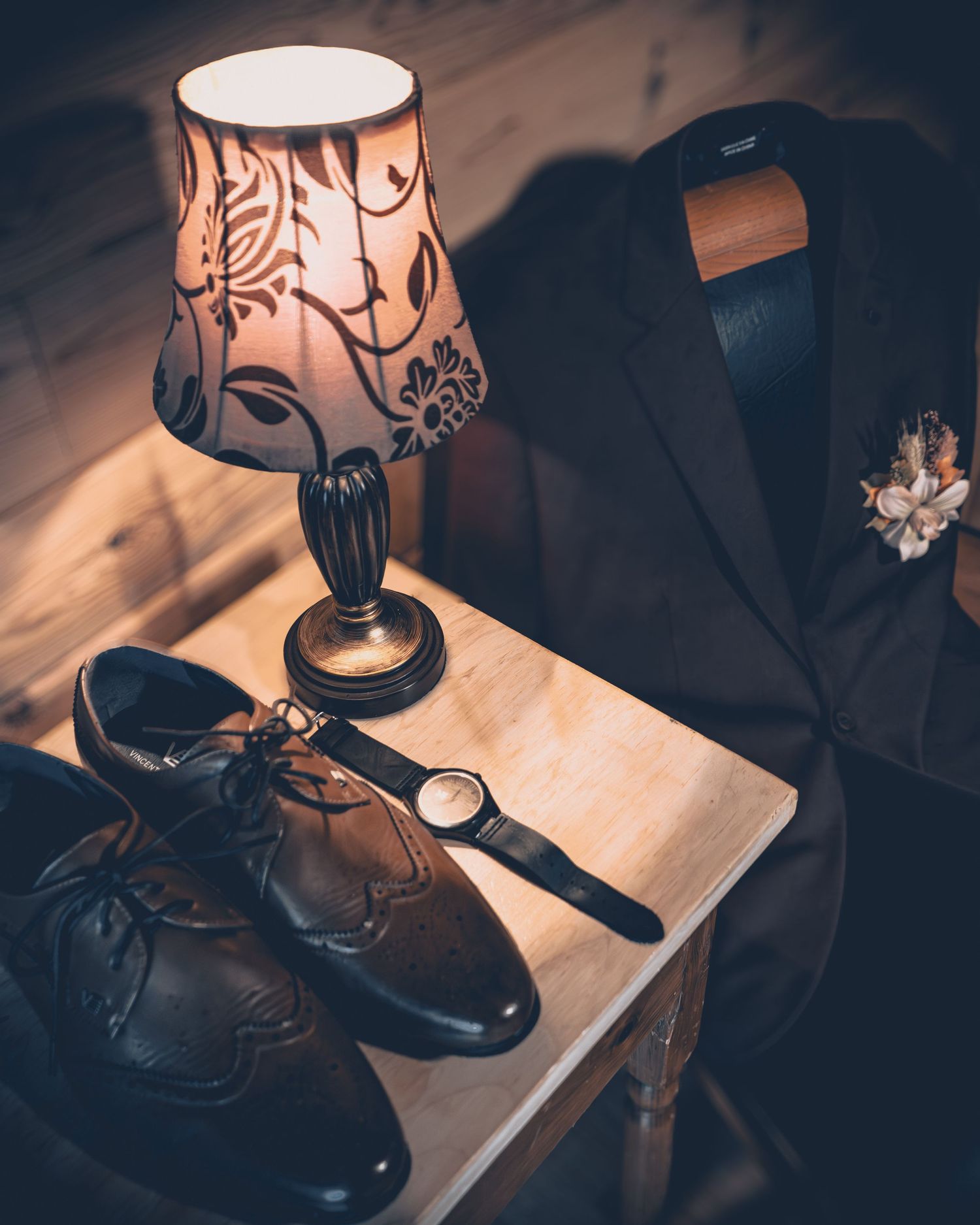 A vintage table lamp illuminates dress shoes and a suit jacket in a moody wedding preparation setting.