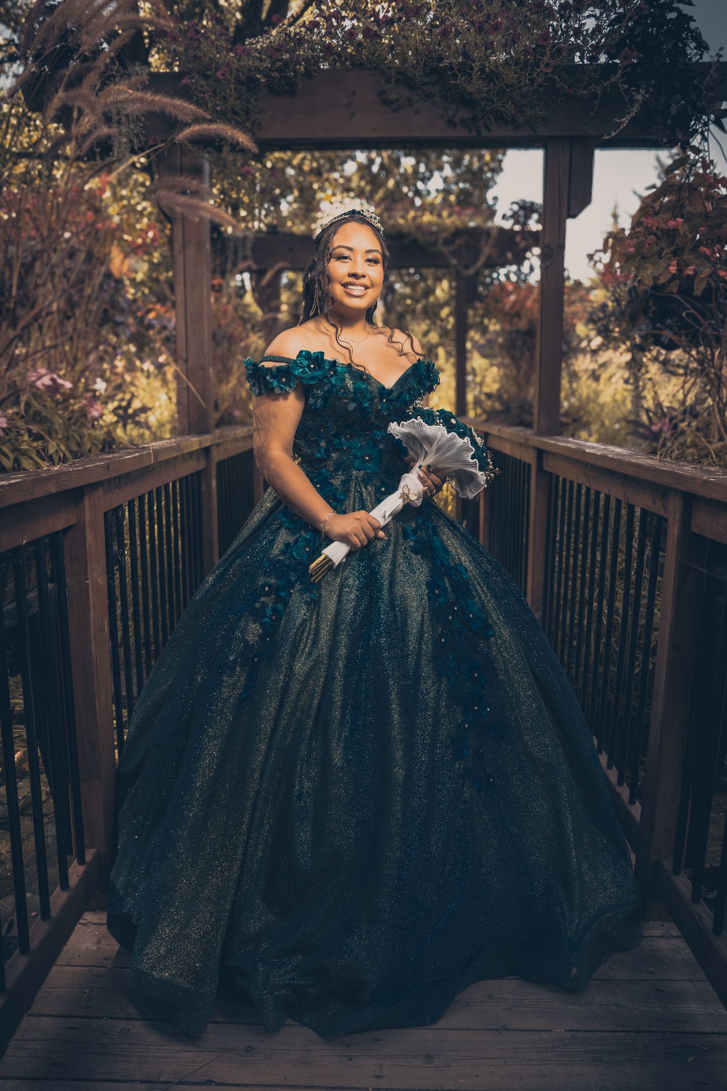 Person in a teal ball gown poses on a wooden deck surrounded by autumn foliage.