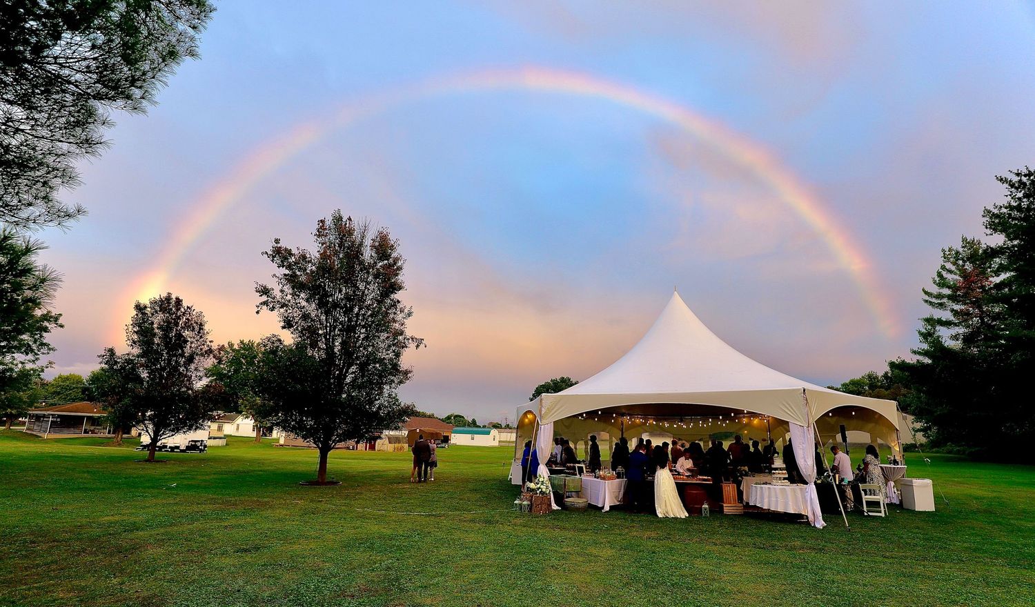 Stunning double rainbow arching over white wedding reception tent on lush green lawn at dusk.