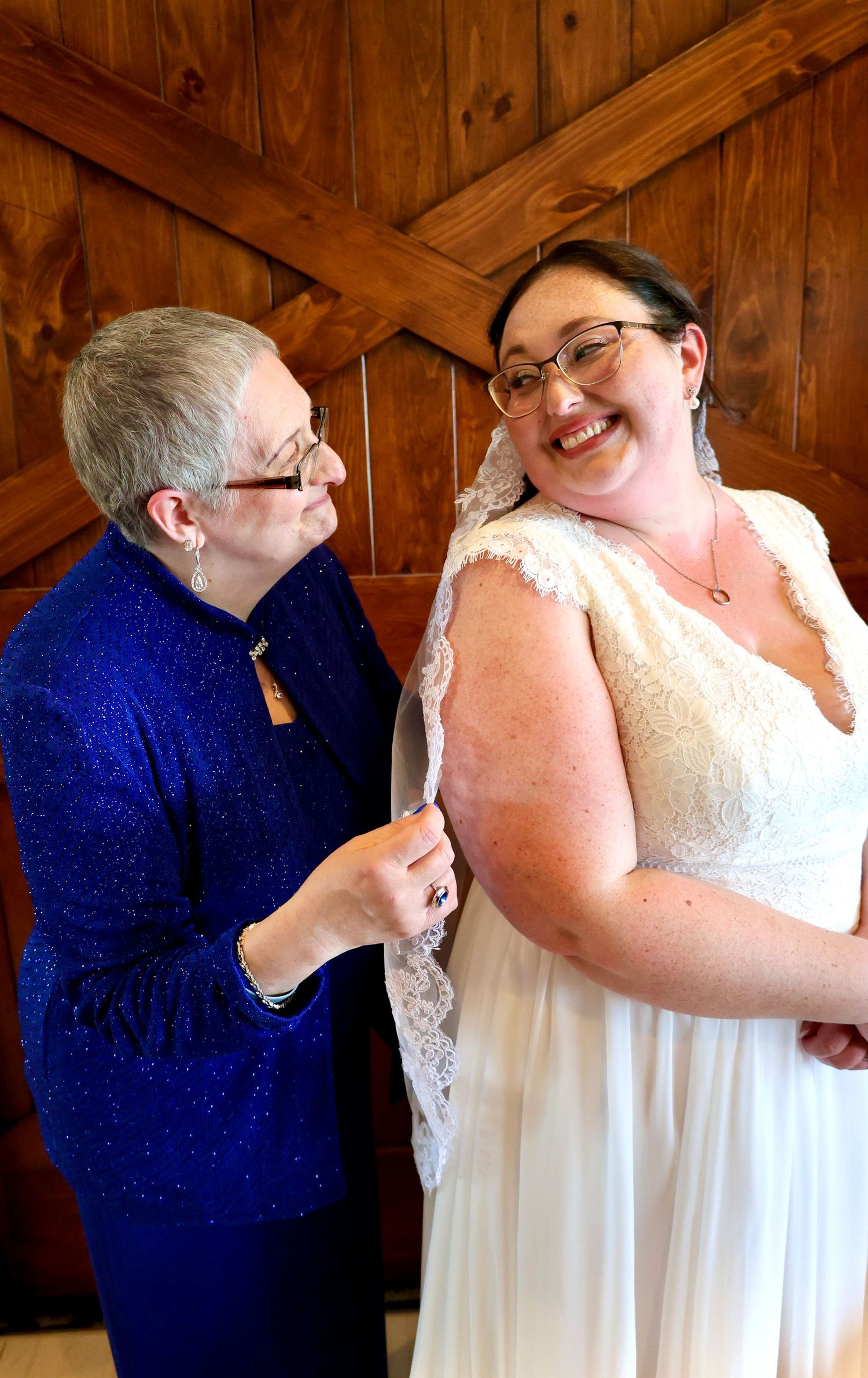 Older woman in blue jacket helping a smiling bride adjust her lace wedding dress in a barn venue.