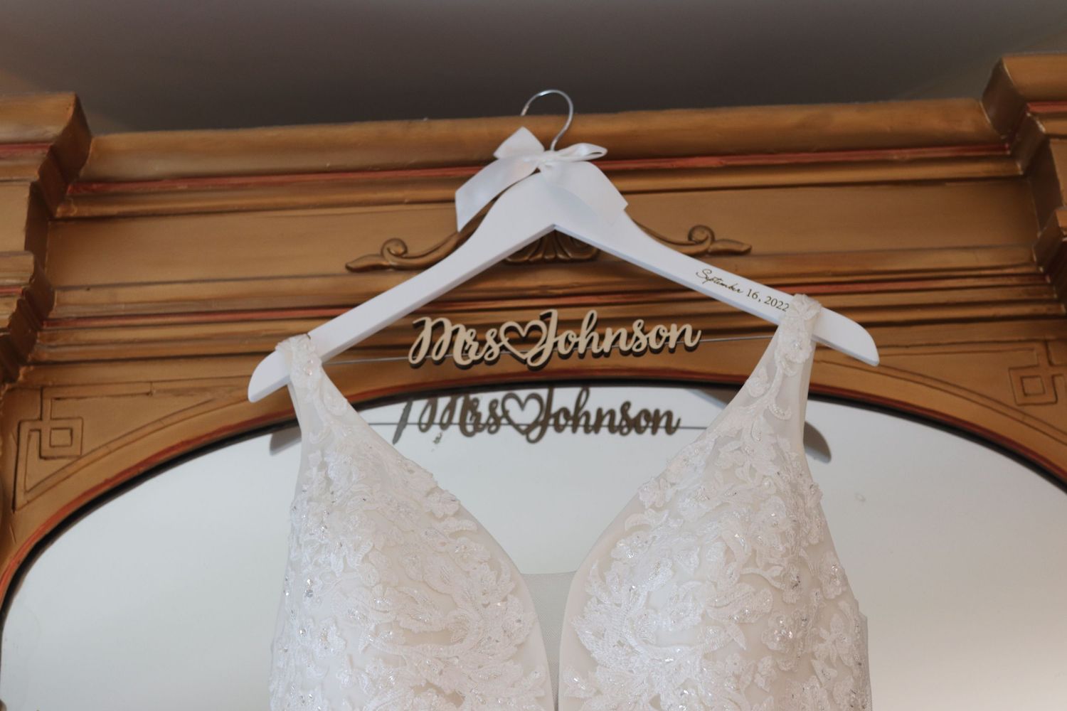 White lace wedding dress hanging on a custom Mrs. Johnson bridal hanger on a wooden mirror.