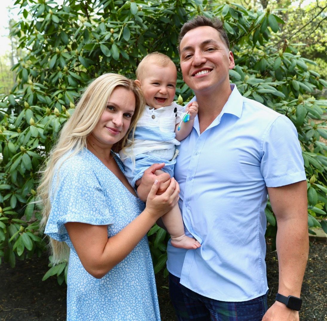 Happy family with blonde mother, smiling father, and laughing baby boy posing outdoors.