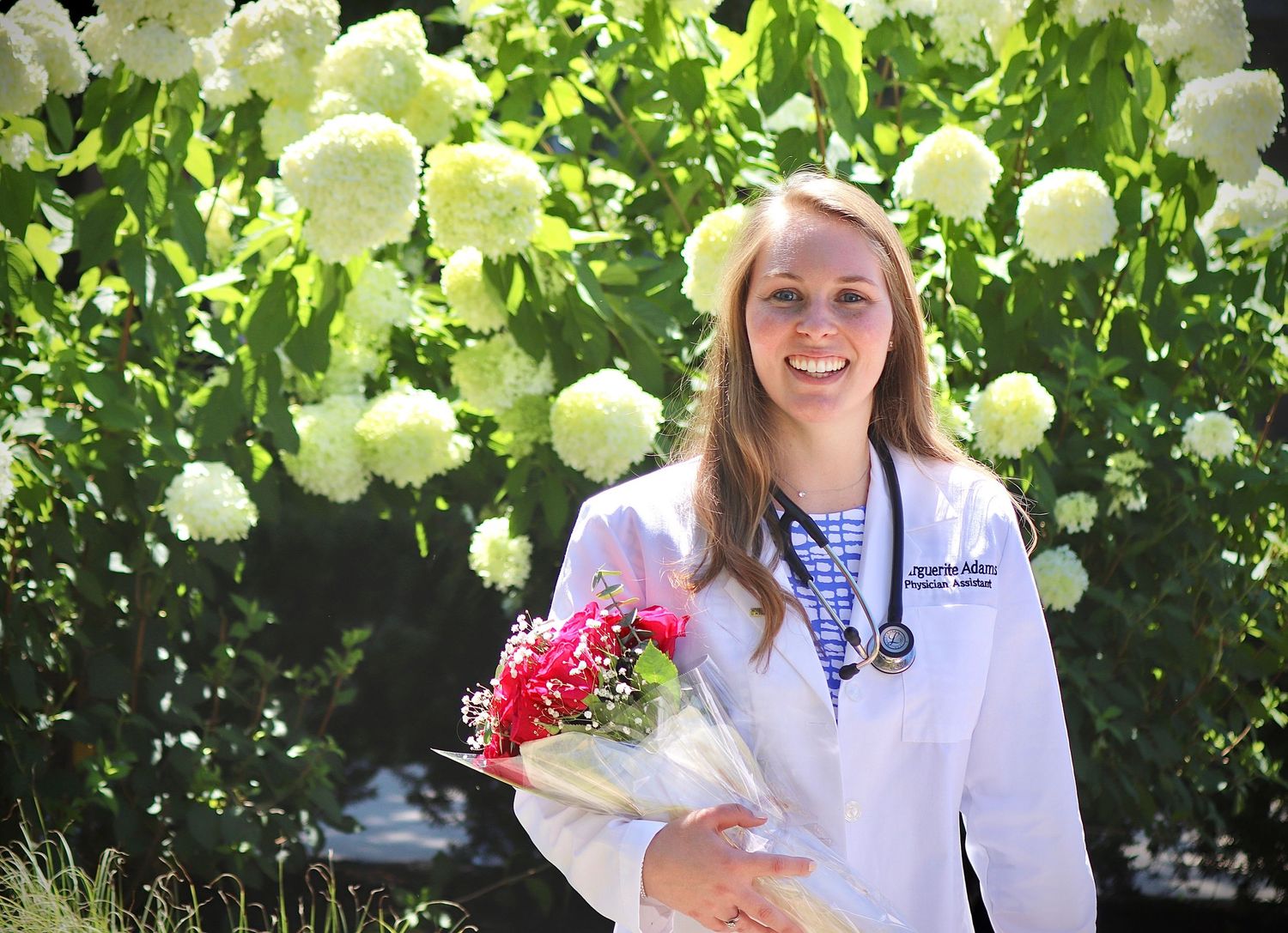 Young woman in white lab coat holding red flowers poses outdoors near blooming white hydrangeas.