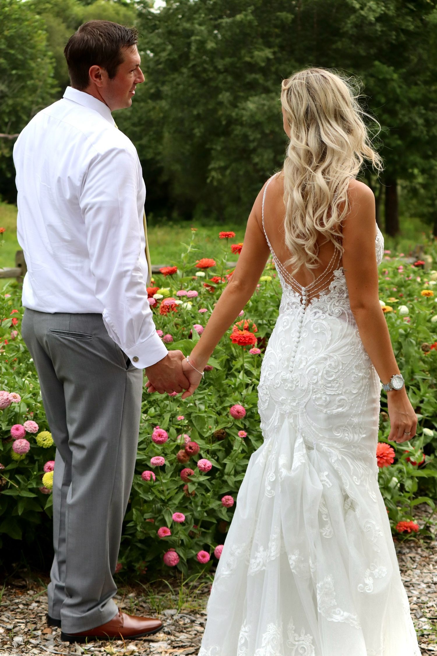 Bride in backless lace mermaid gown holds hands with groom in white shirt walking through colorful flower garden.