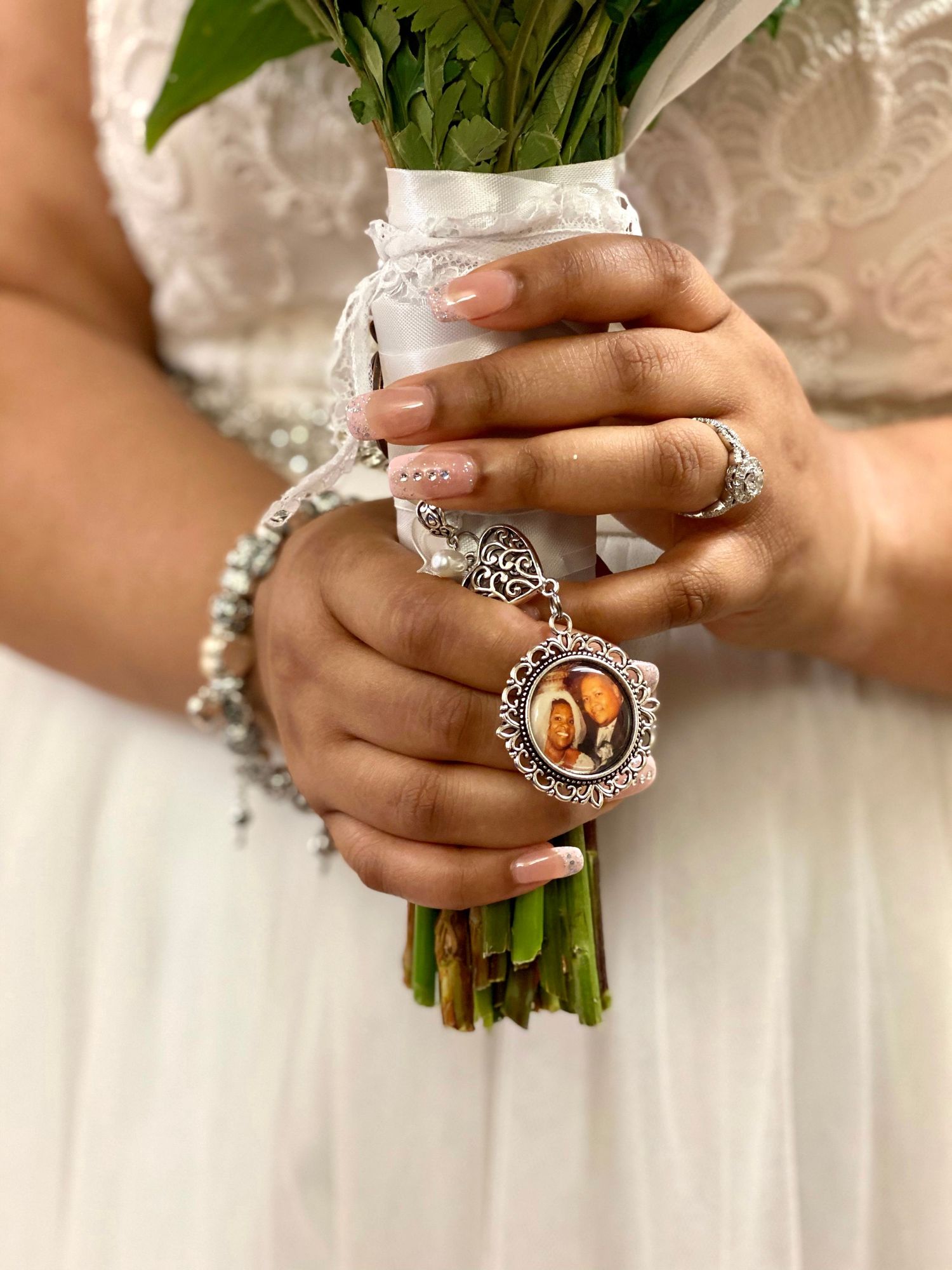 Bride holding white bouquet with memorial photo charm, wearing pearl bracelet and rings, in white wedding dress.