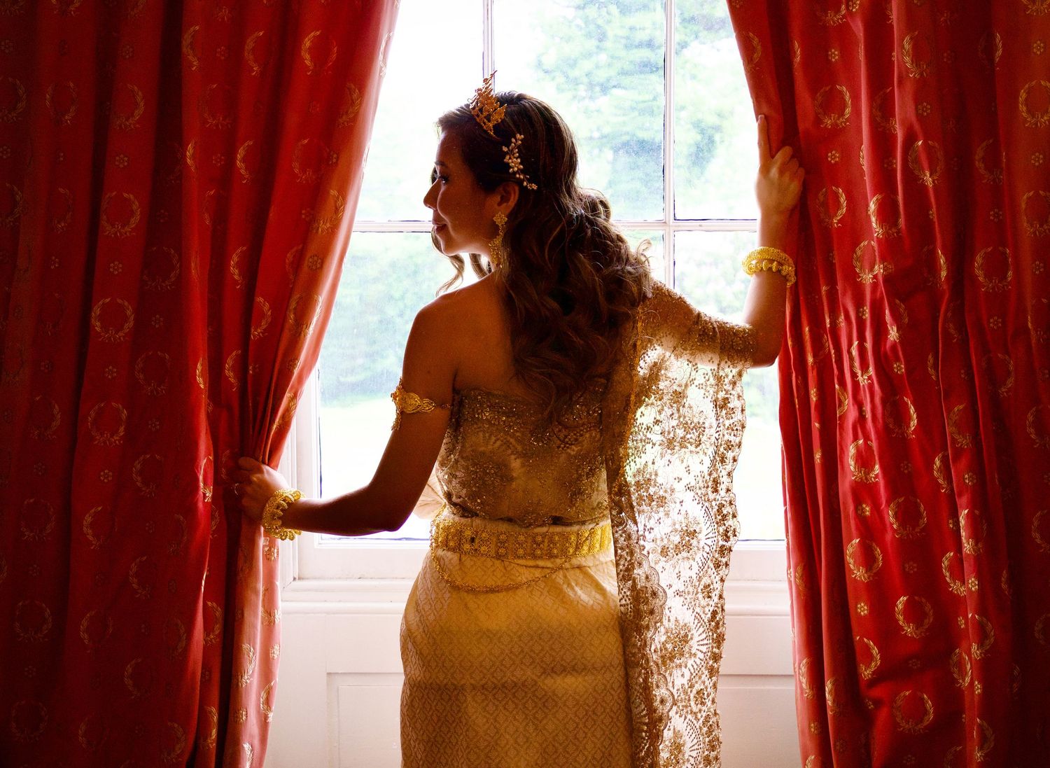 Elegant bride in gold lace gown standing backlit by window framed by red ornate curtains.