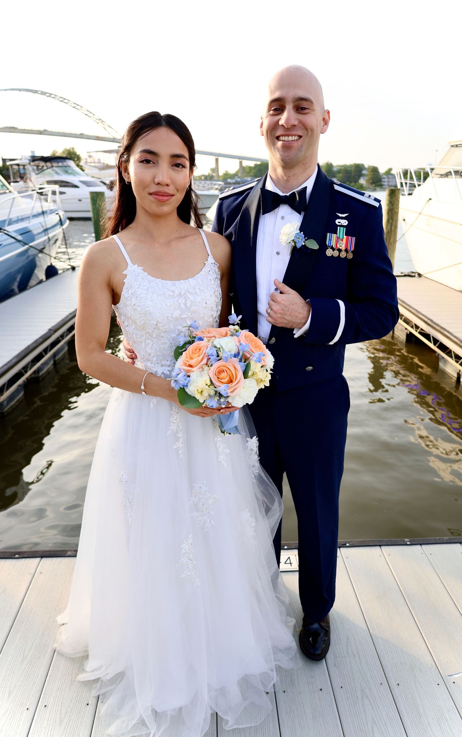 Bride and groom in military dress uniform pose together on marina dock at sunset.