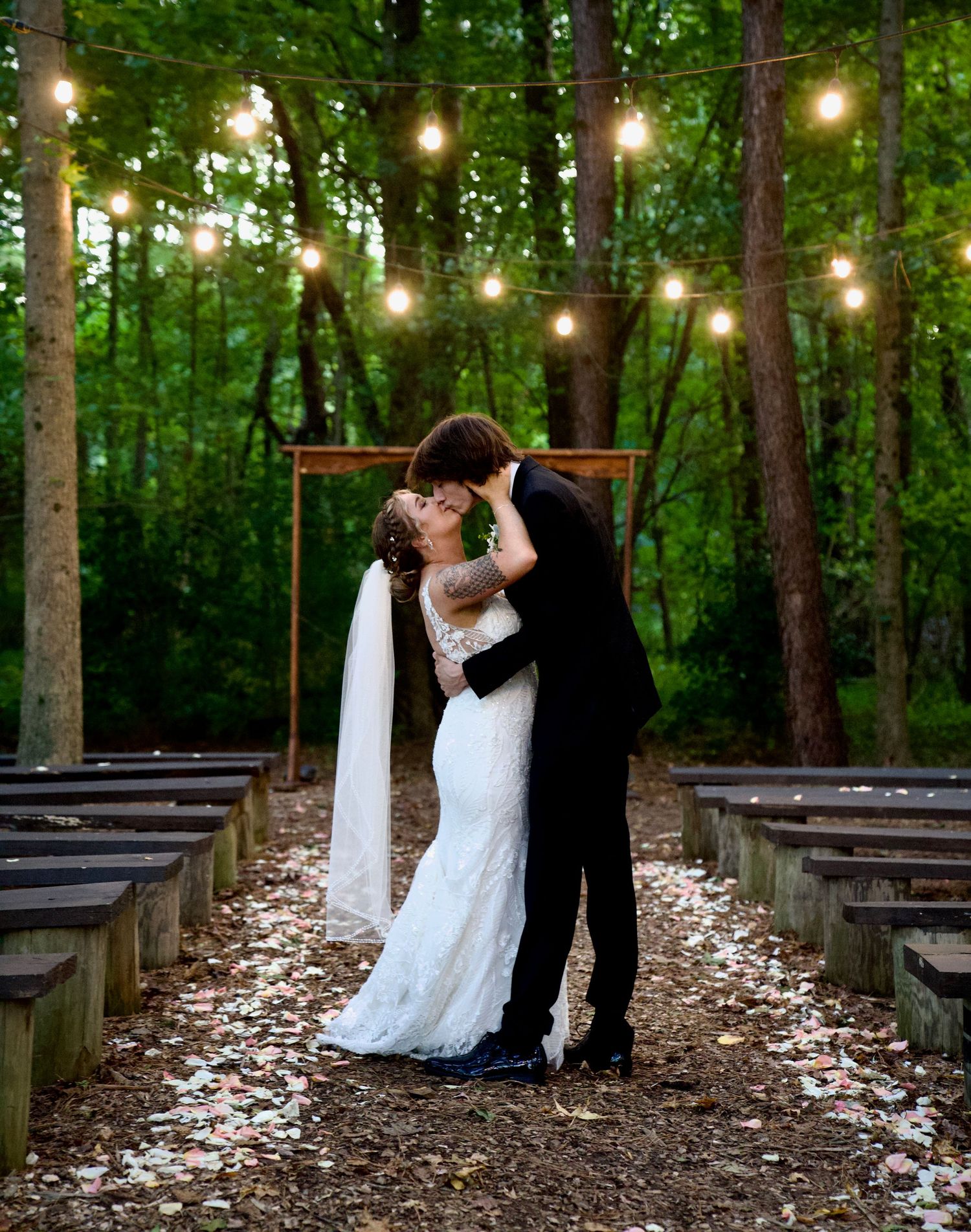 Bride and groom kissing at an enchanting outdoor forest wedding ceremony with string lights.