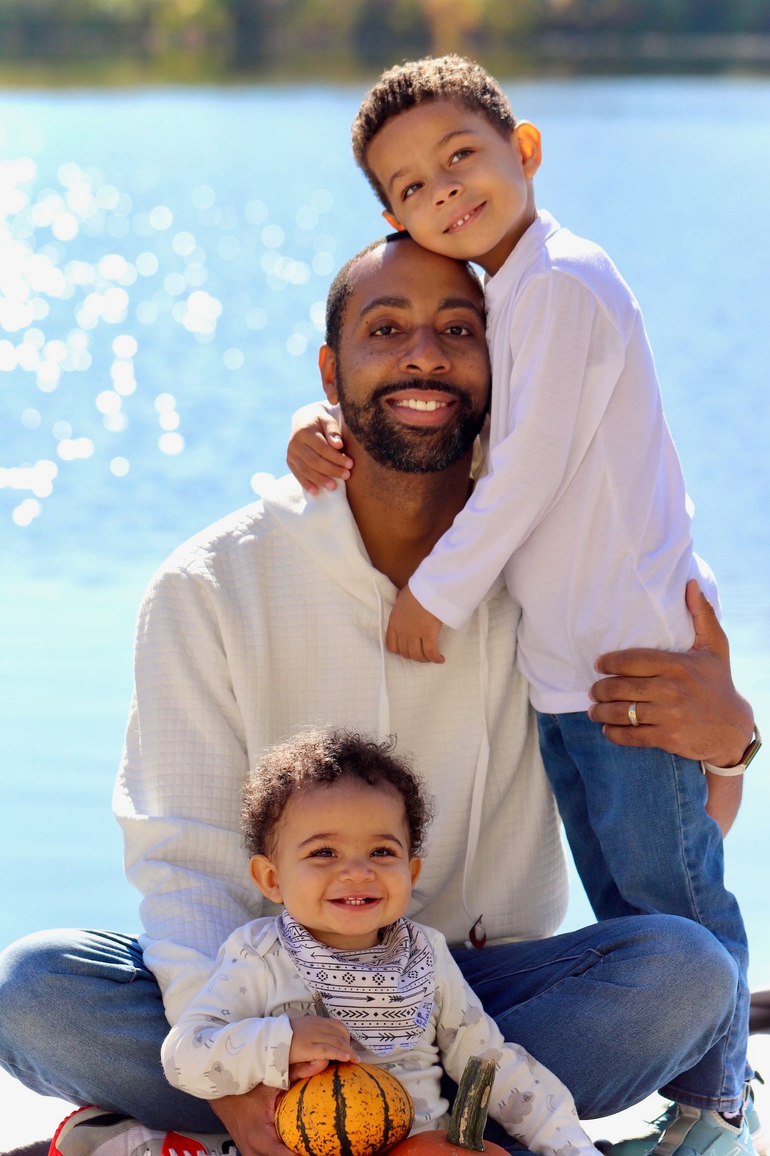 Smiling father with two young sons posing outdoors by a bright sunlit lake.