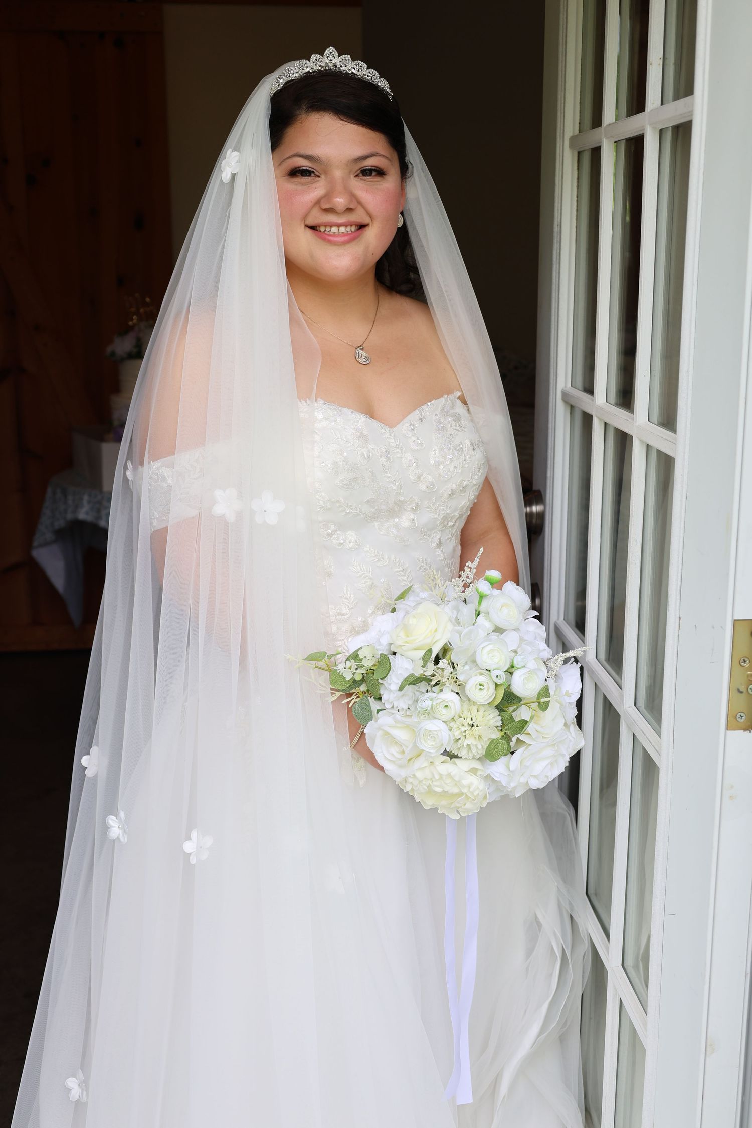 Smiling bride in strapless lace gown with tiara, veil, and white rose bouquet standing in doorway.