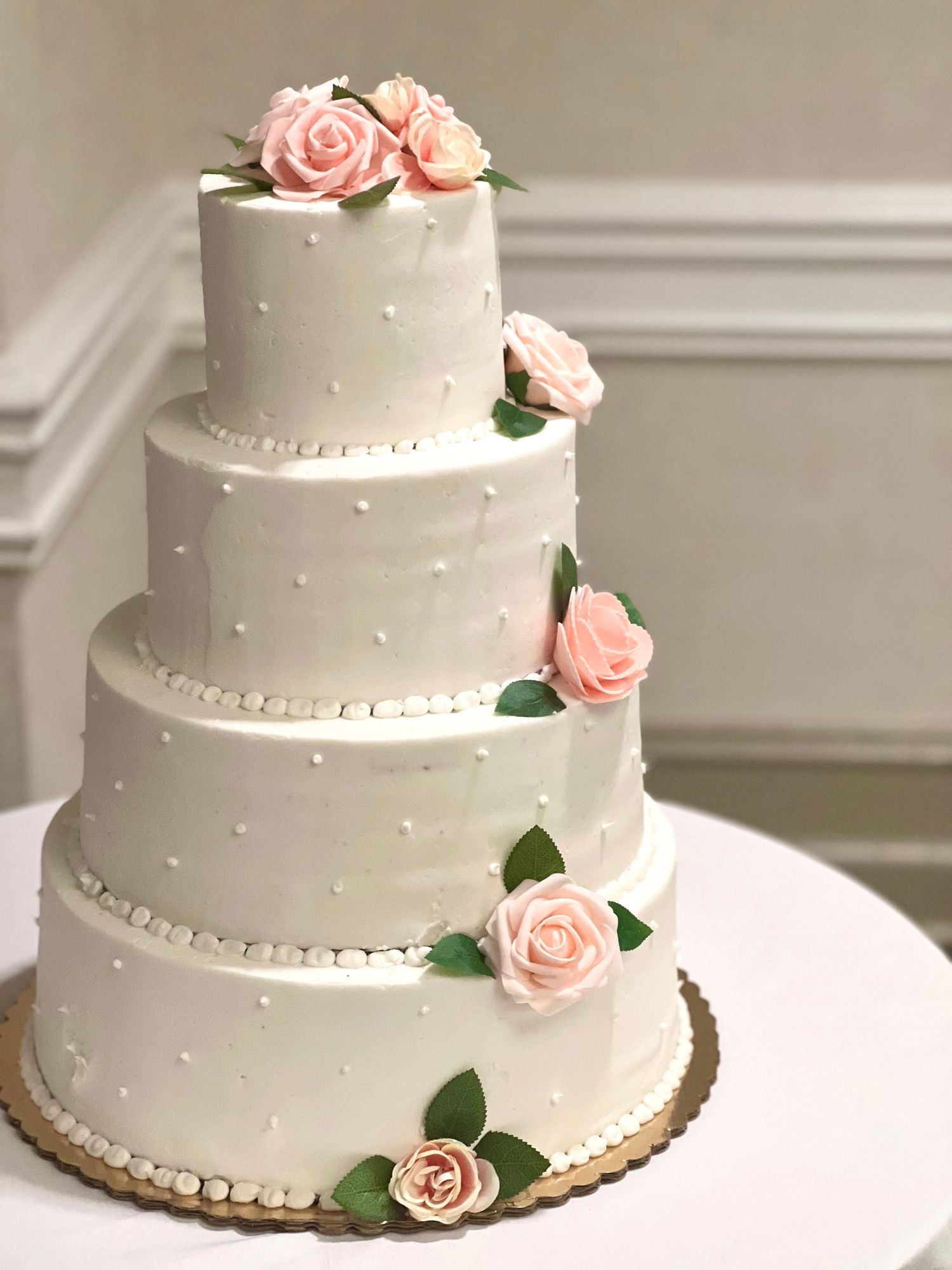 Elegant four-tier white wedding cake decorated with pink roses and pearl dot details on white stand.