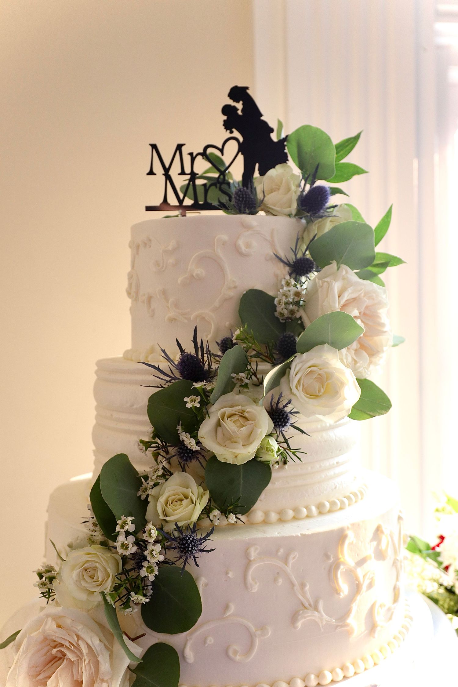 Three-tier white wedding cake decorated with roses, eucalyptus, and Mr. and Mrs. silhouette cake topper.