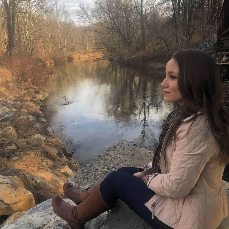 Young woman in pink jacket sits on rocks gazing at peaceful creek surrounded by bare autumn trees.