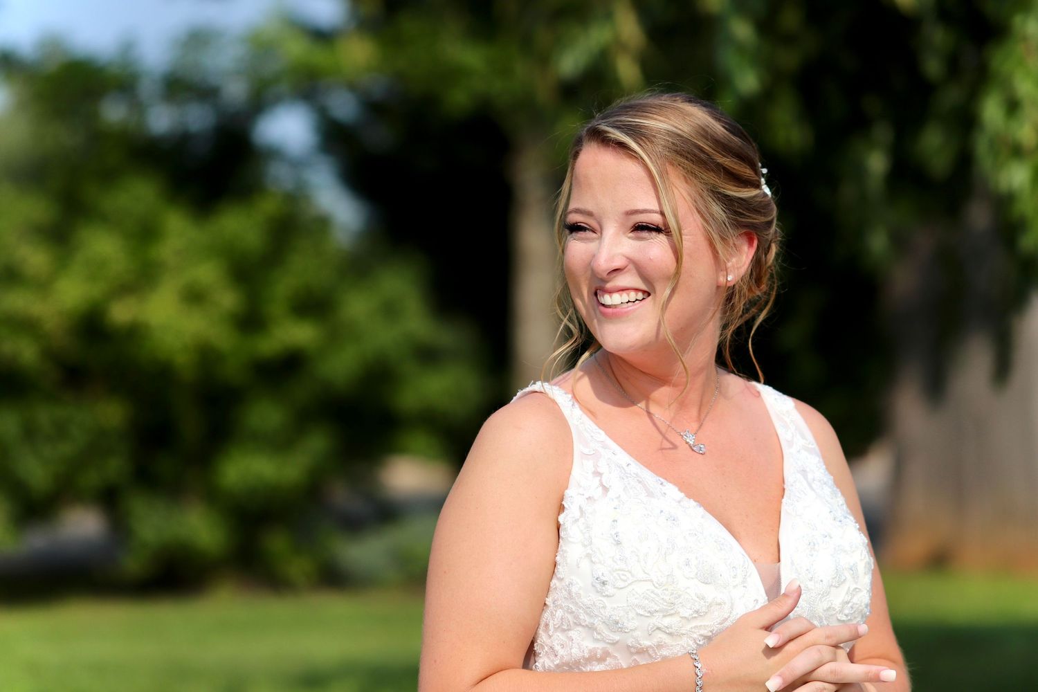 Smiling bride in white lace wedding gown standing outdoors with lush green trees in background.