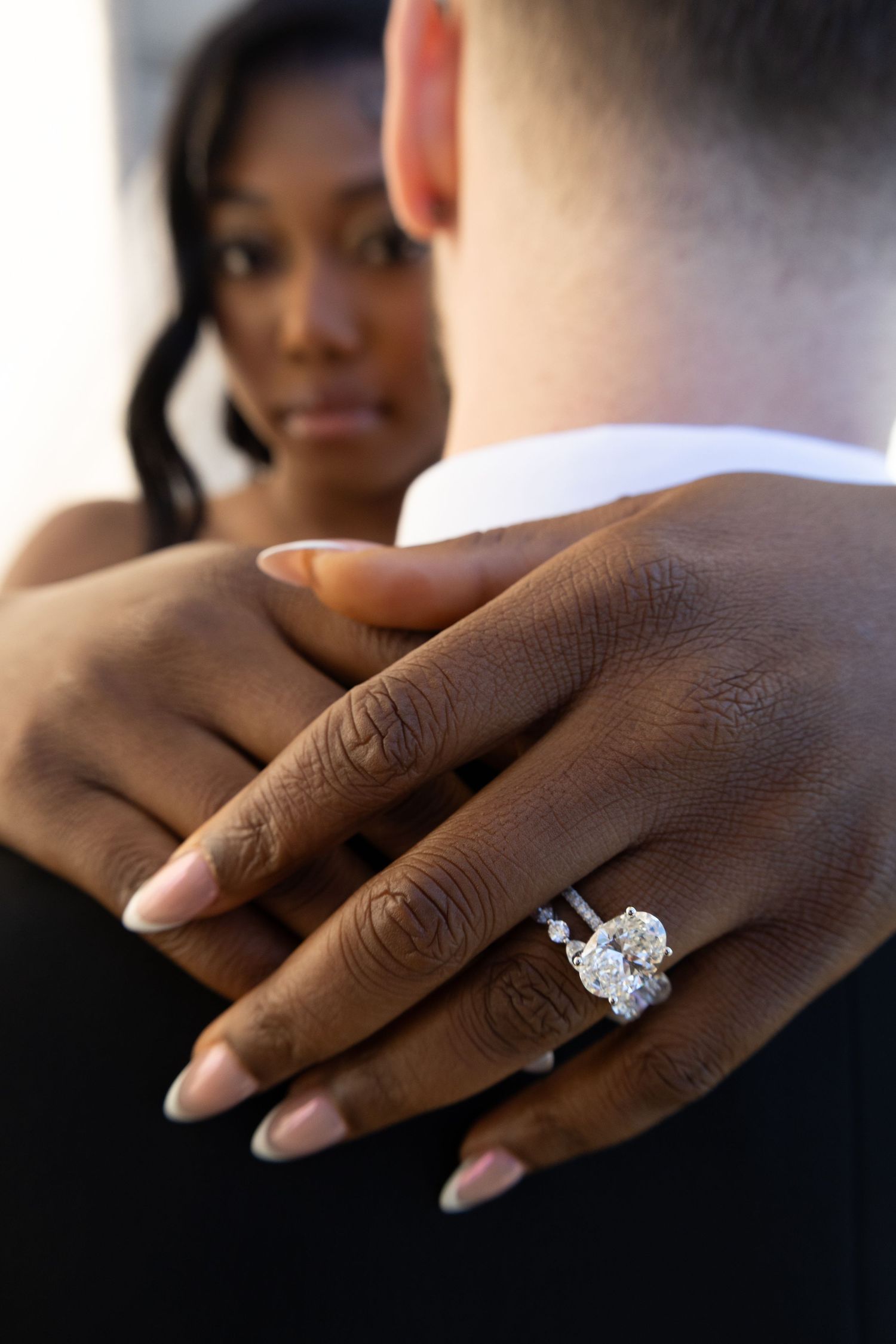 Close-up of a stunning oval diamond engagement ring on a woman's hand resting on her partner's shoulder.