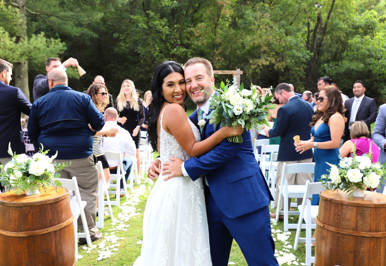 Happy newlywed couple embracing and smiling at outdoor garden wedding ceremony with guests behind.