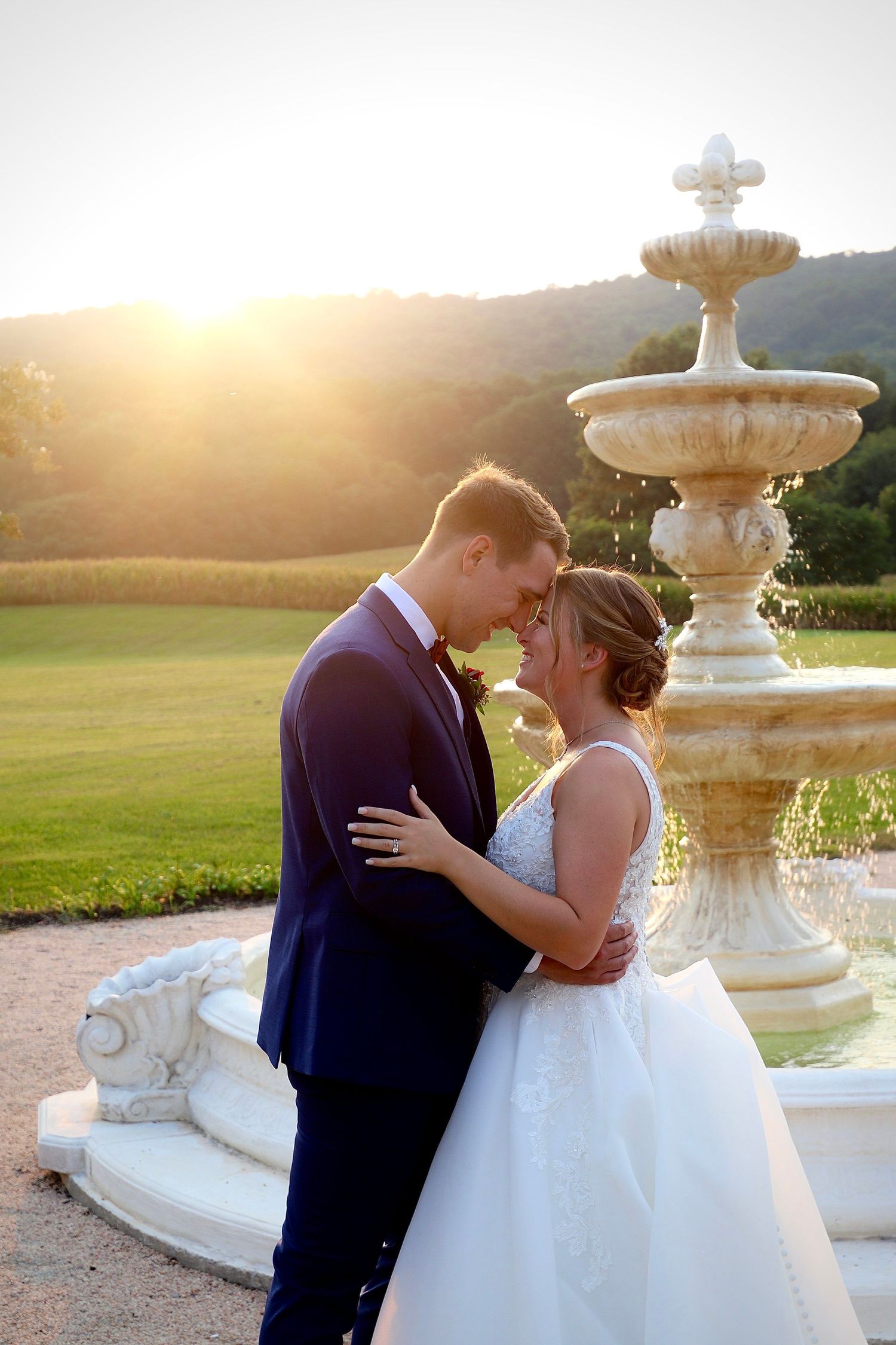 Couple sharing romantic moment by stone fountain at golden hour sunset with rolling green hills backdrop.
