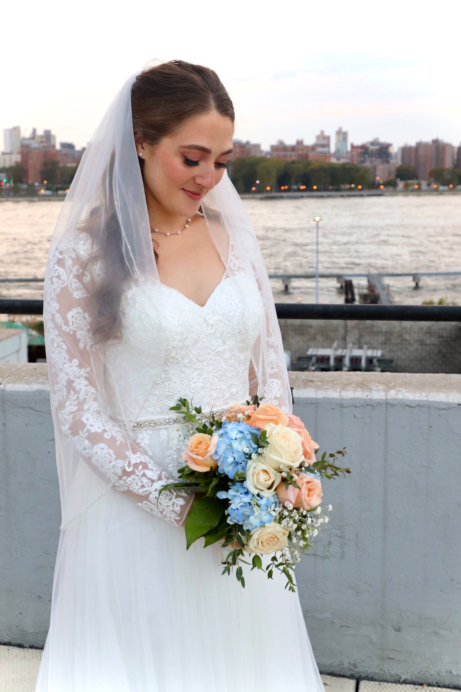 Beautiful bride in lace gown with veil holds colorful bouquet by waterfront at dusk.