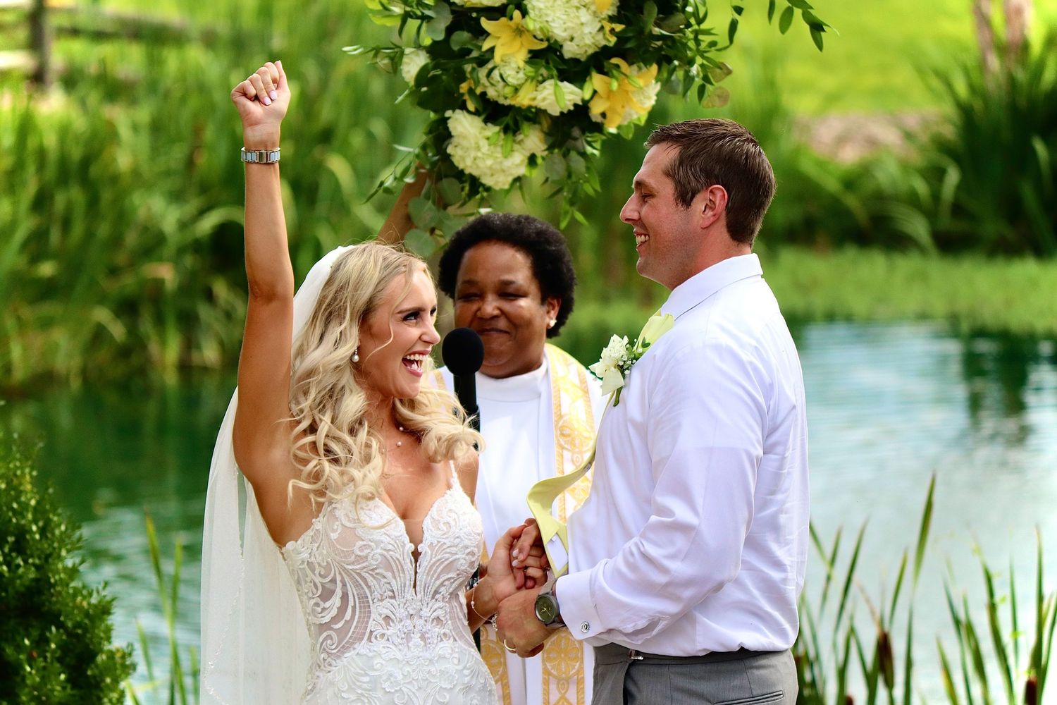 Joyful bride raises fist at outdoor lakeside wedding ceremony with groom and officiant.