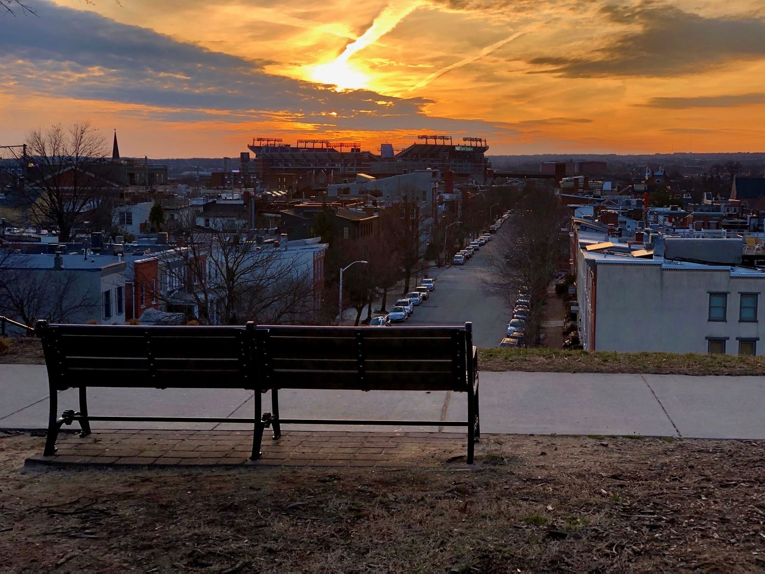 Park bench overlooking city skyline at sunset with stadium visible in the golden distance.