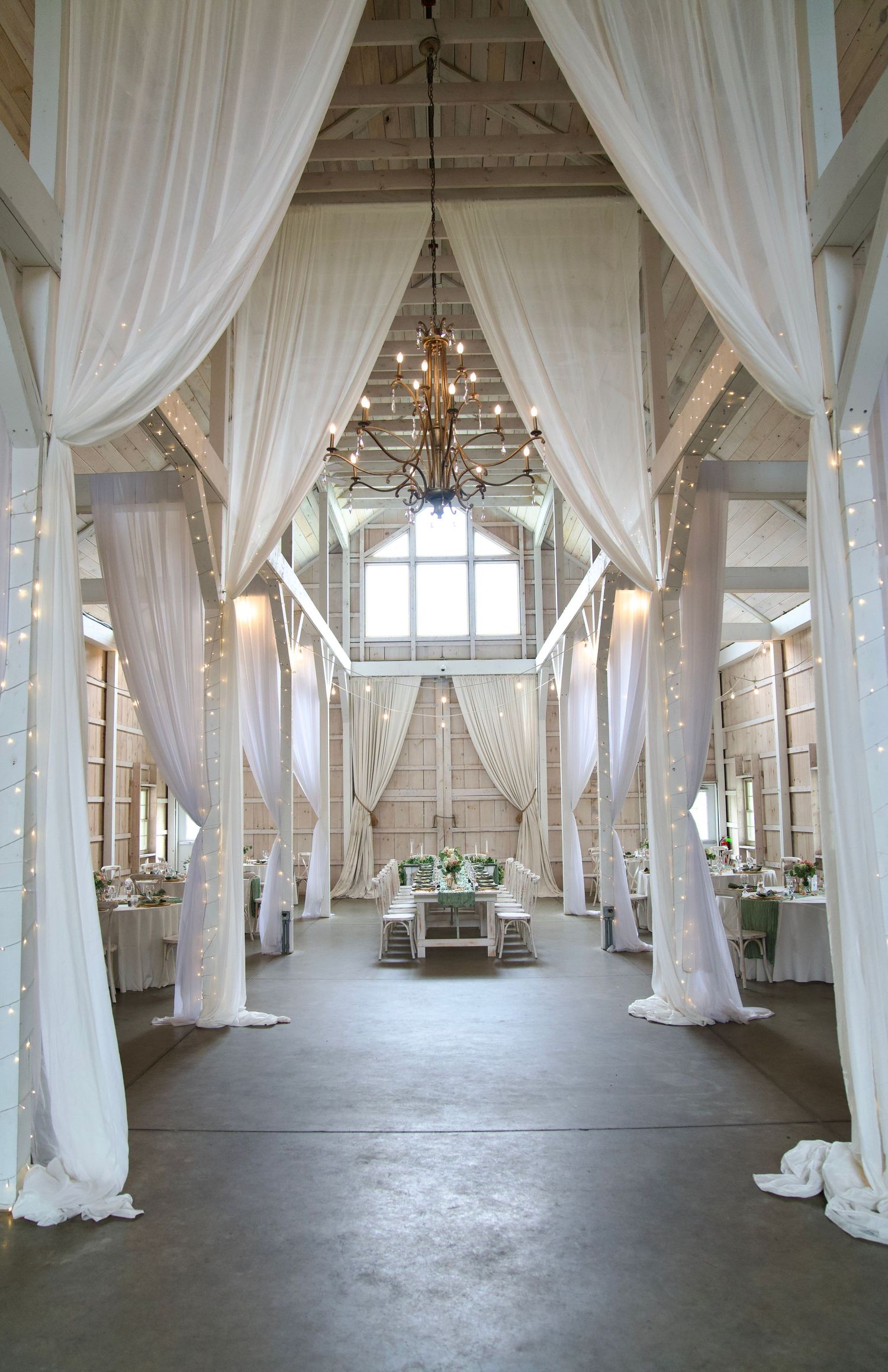 Stunning barn wedding venue with white draping columns chandelier and string lights decor.
