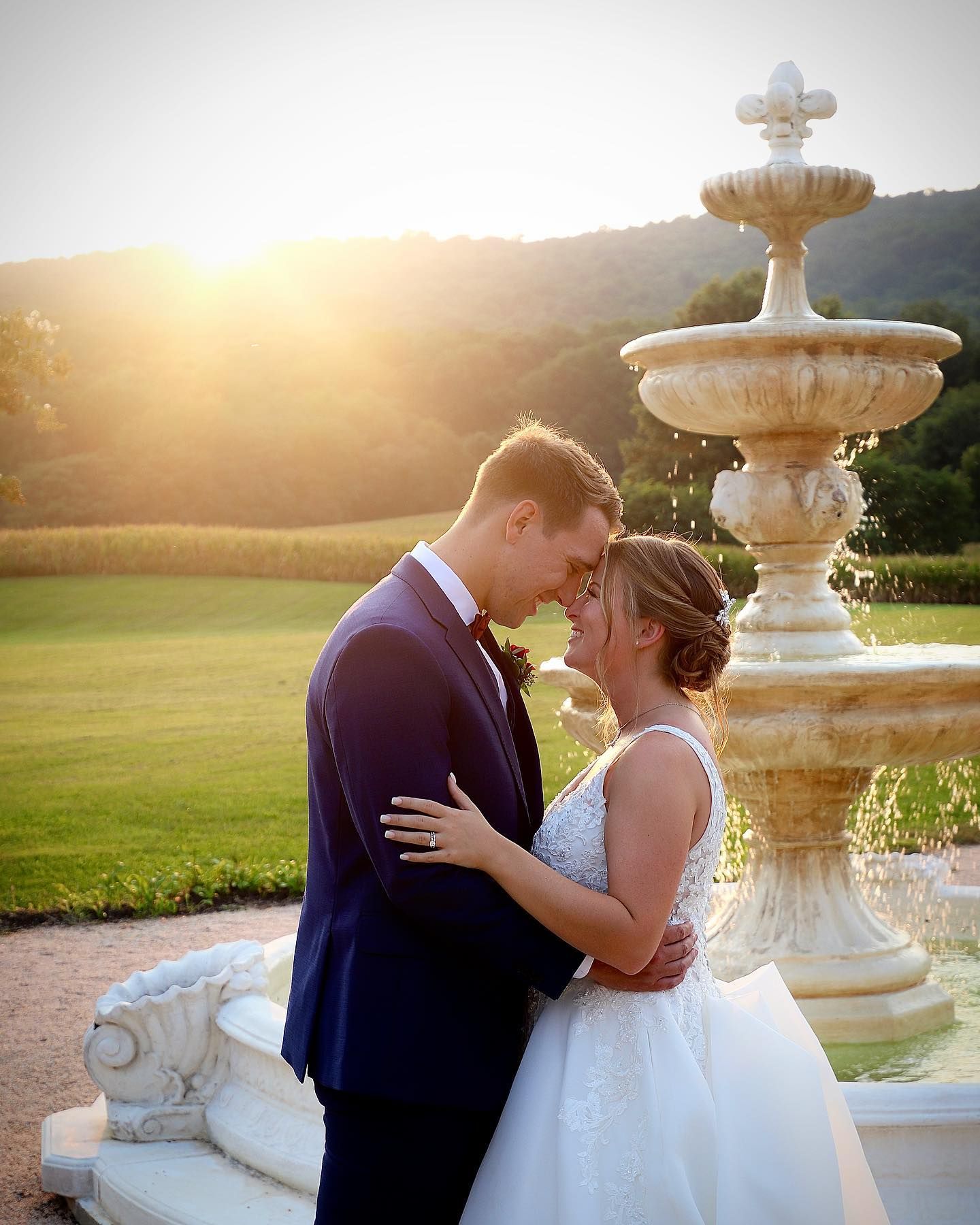 A romantic sunset wedding photo by an ornate white fountain in a garden setting.