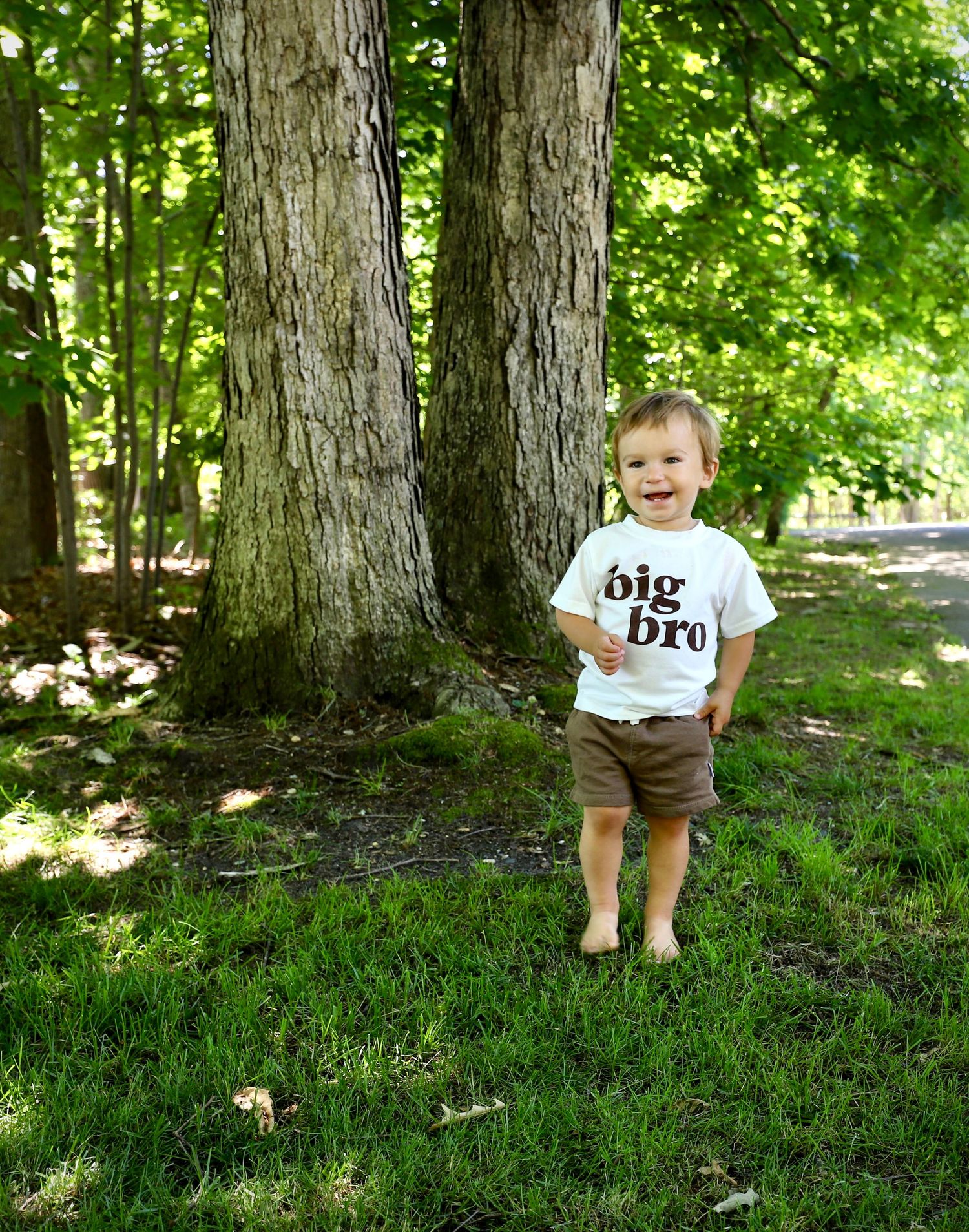 A toddler in a 'big bro' t-shirt stands smiling between two large trees in a sunny park.