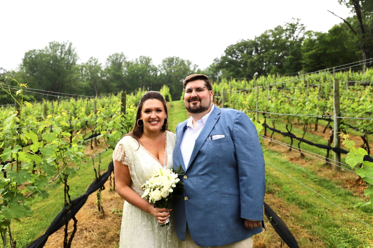 Smiling newlywed couple posing together with white bouquet in a lush green vineyard setting.