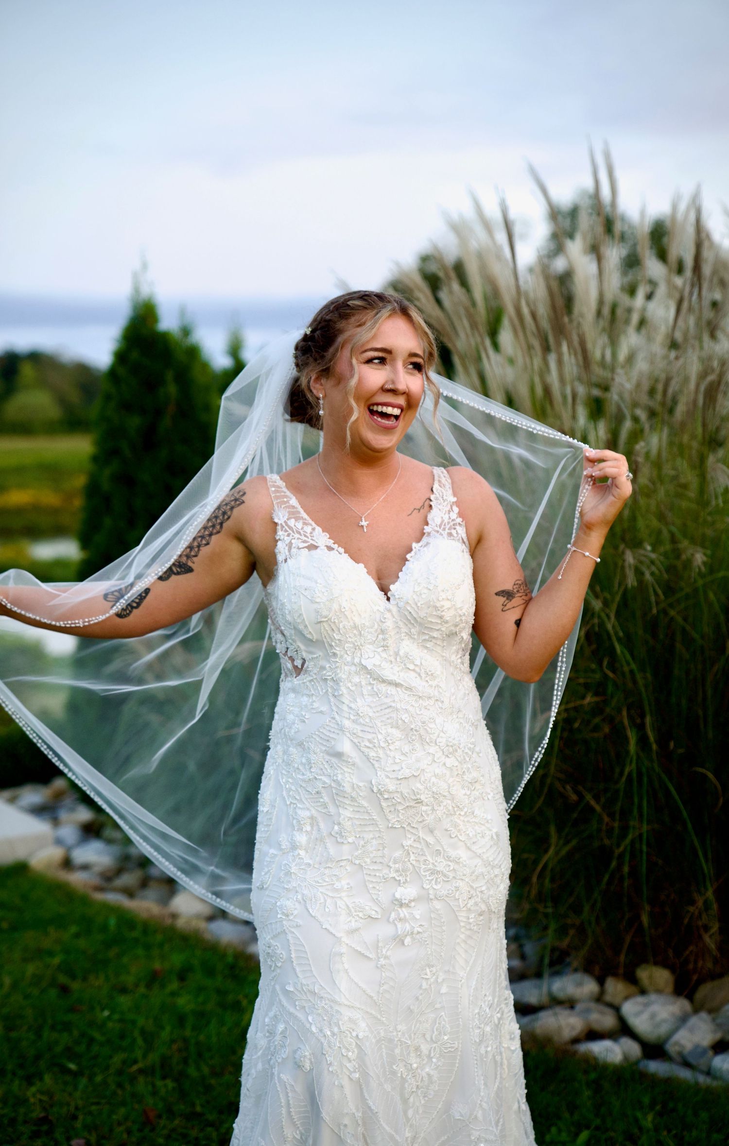 Joyful bride in lace gown holding her veil in the wind at an outdoor waterfront wedding.