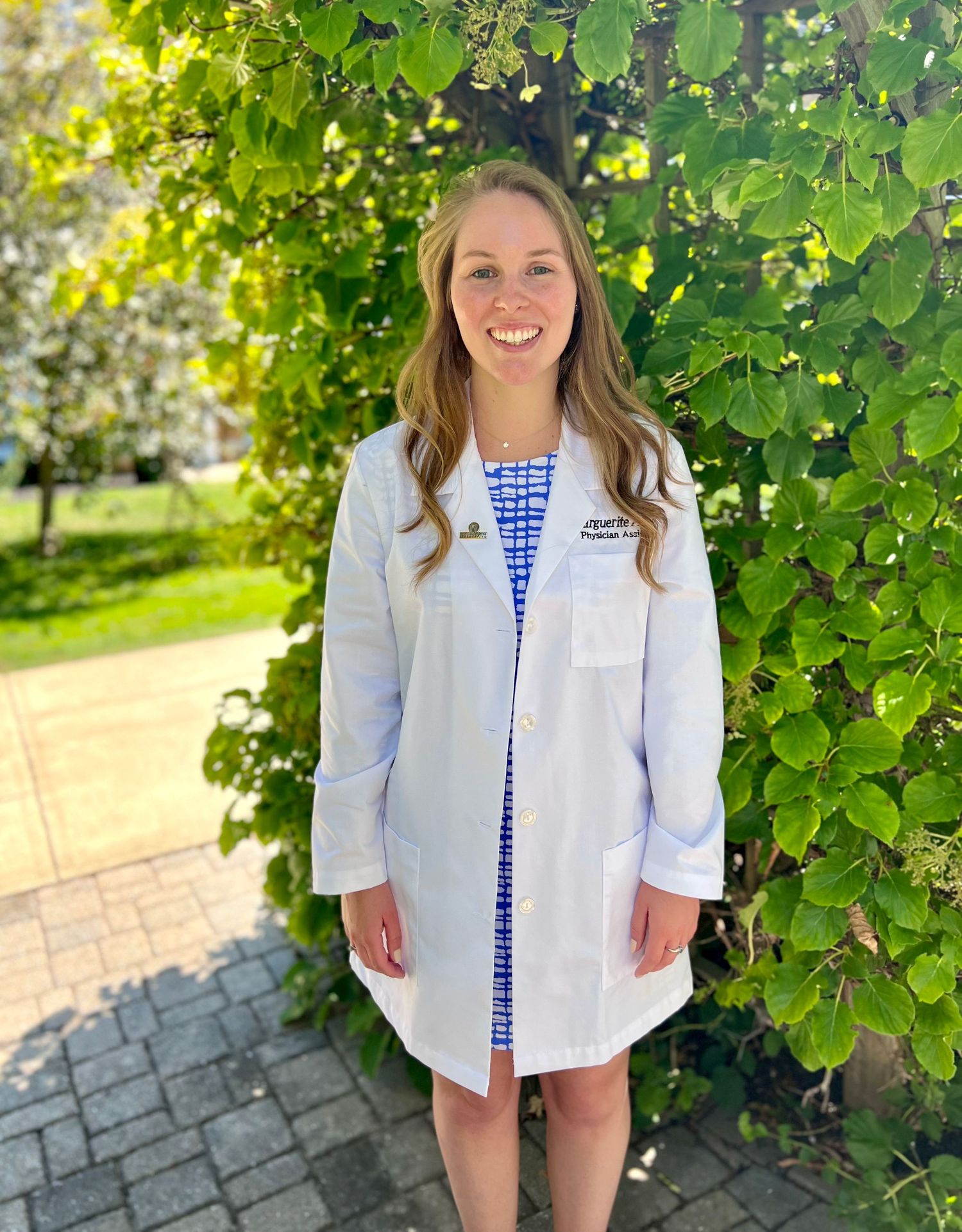 Smiling woman in white medical lab coat stands outdoors beside lush green ivy-covered wall.