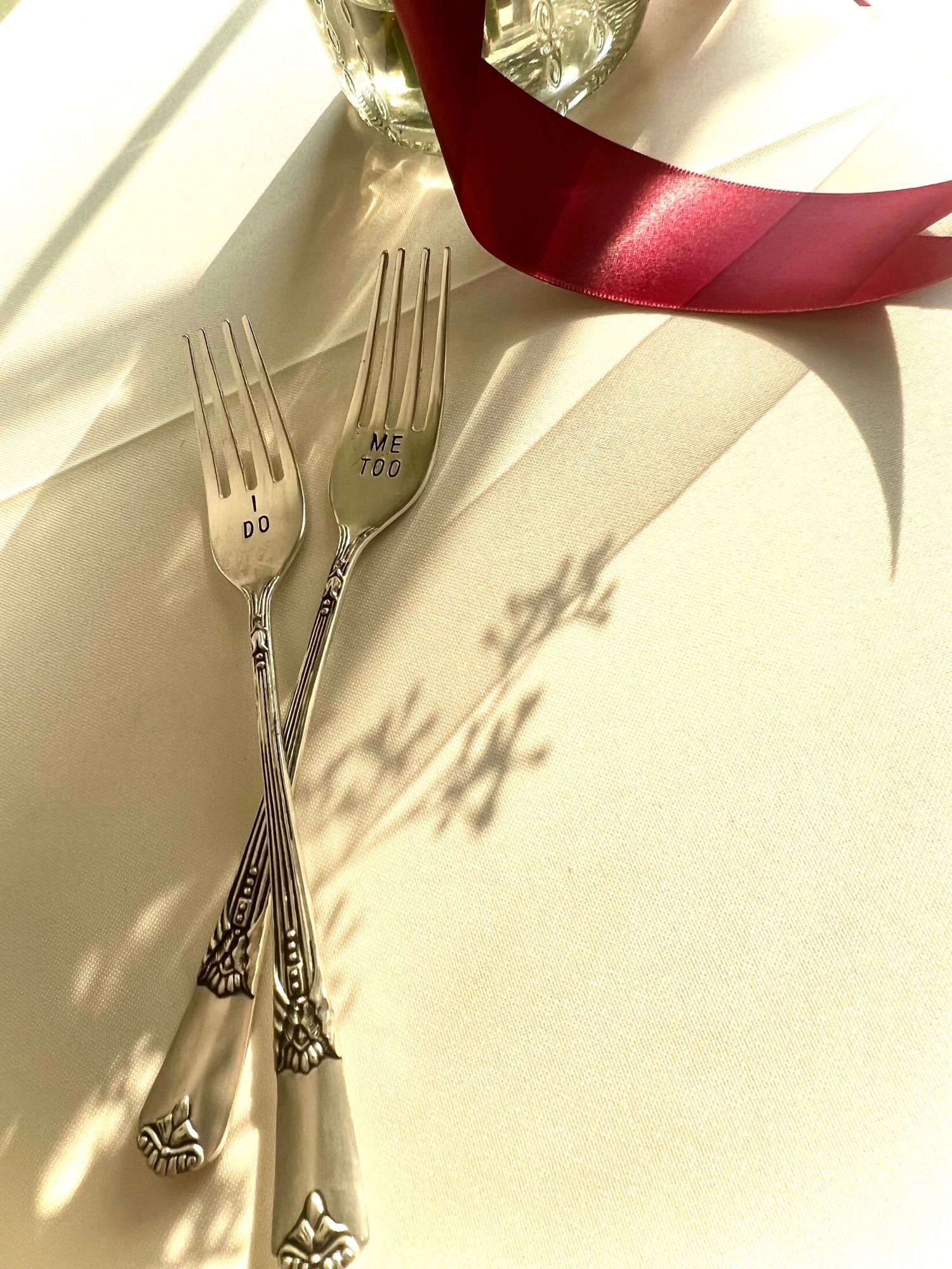 Elegant silver wedding forks engraved with I do and Me too beside a red satin ribbon in sunlight.