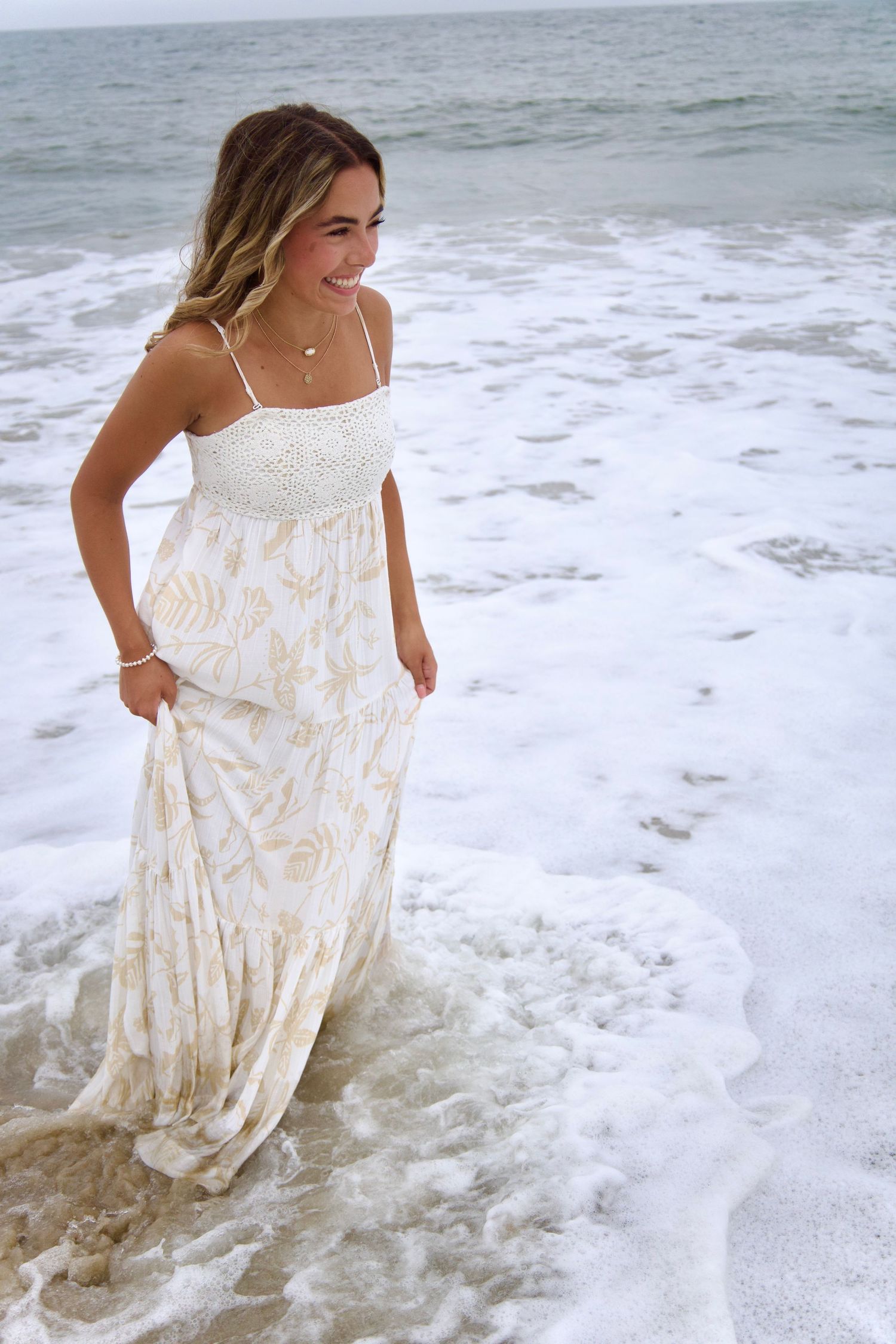 Young woman in floral maxi dress smiling while standing in ocean waves on sandy beach.