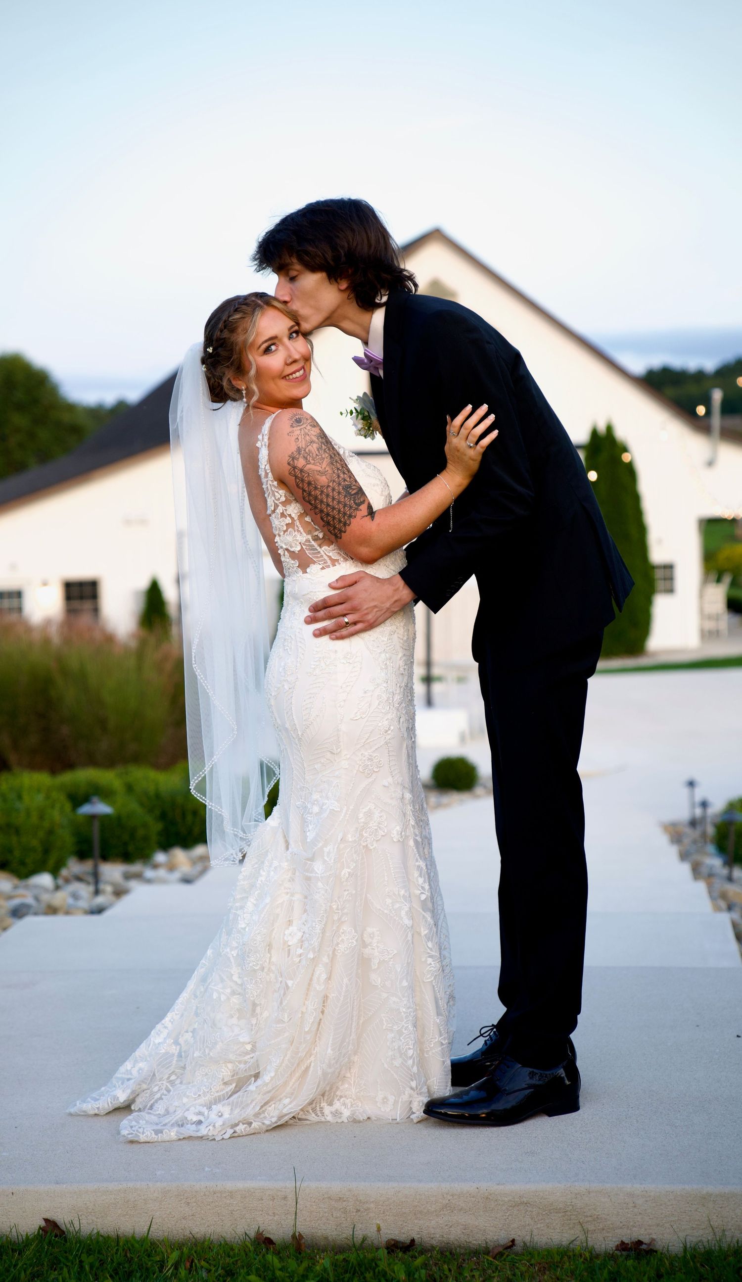 Groom kissing bride in lace mermaid gown with veil outside a white wedding venue at dusk.