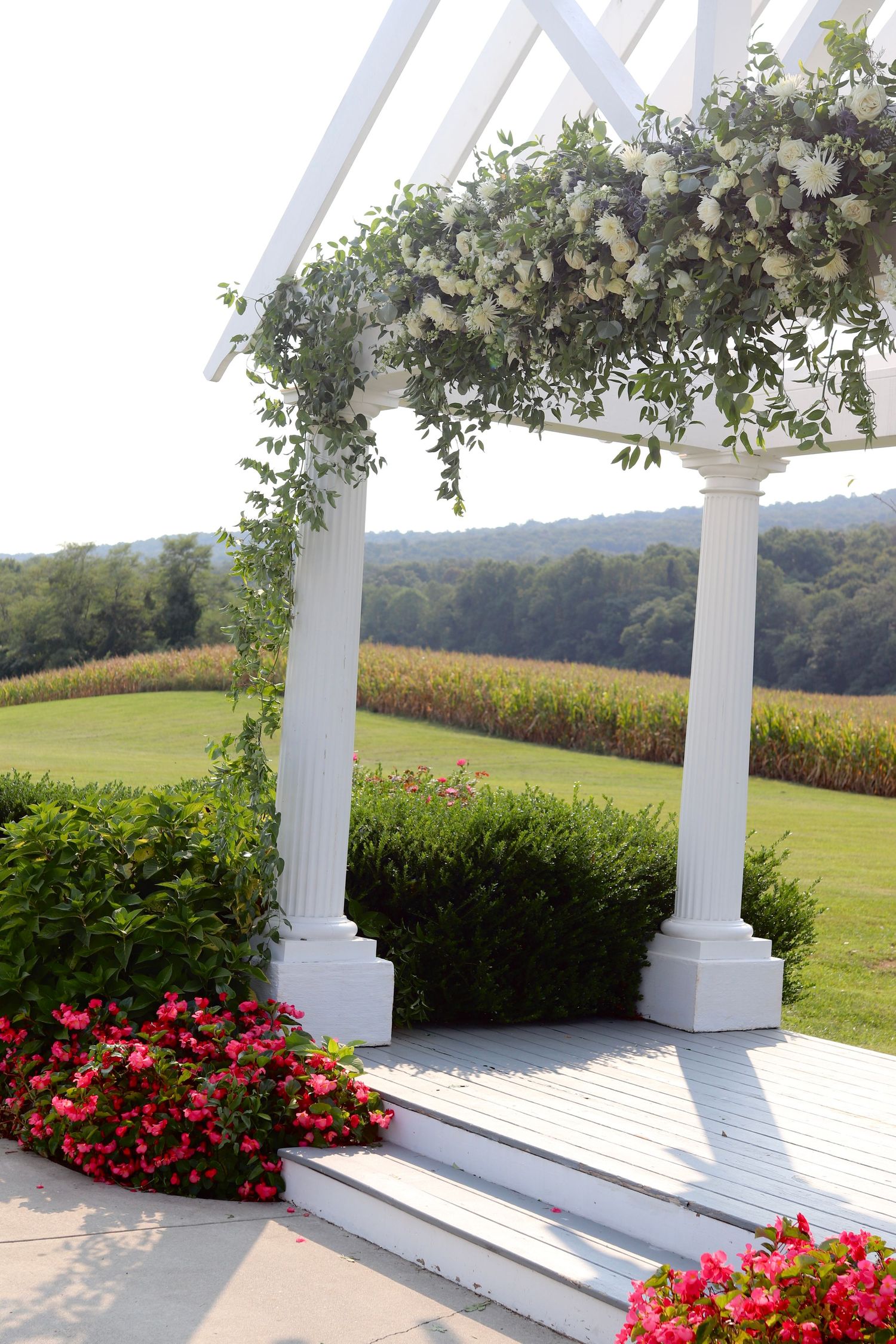 Elegant white wedding pergola adorned with white flowers and greenery overlooking a scenic countryside field.
