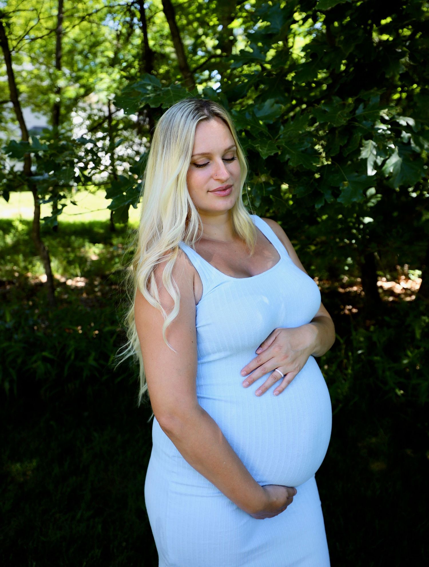 A pregnant woman in a light blue dress cradles her baby bump outdoors surrounded by green foliage.